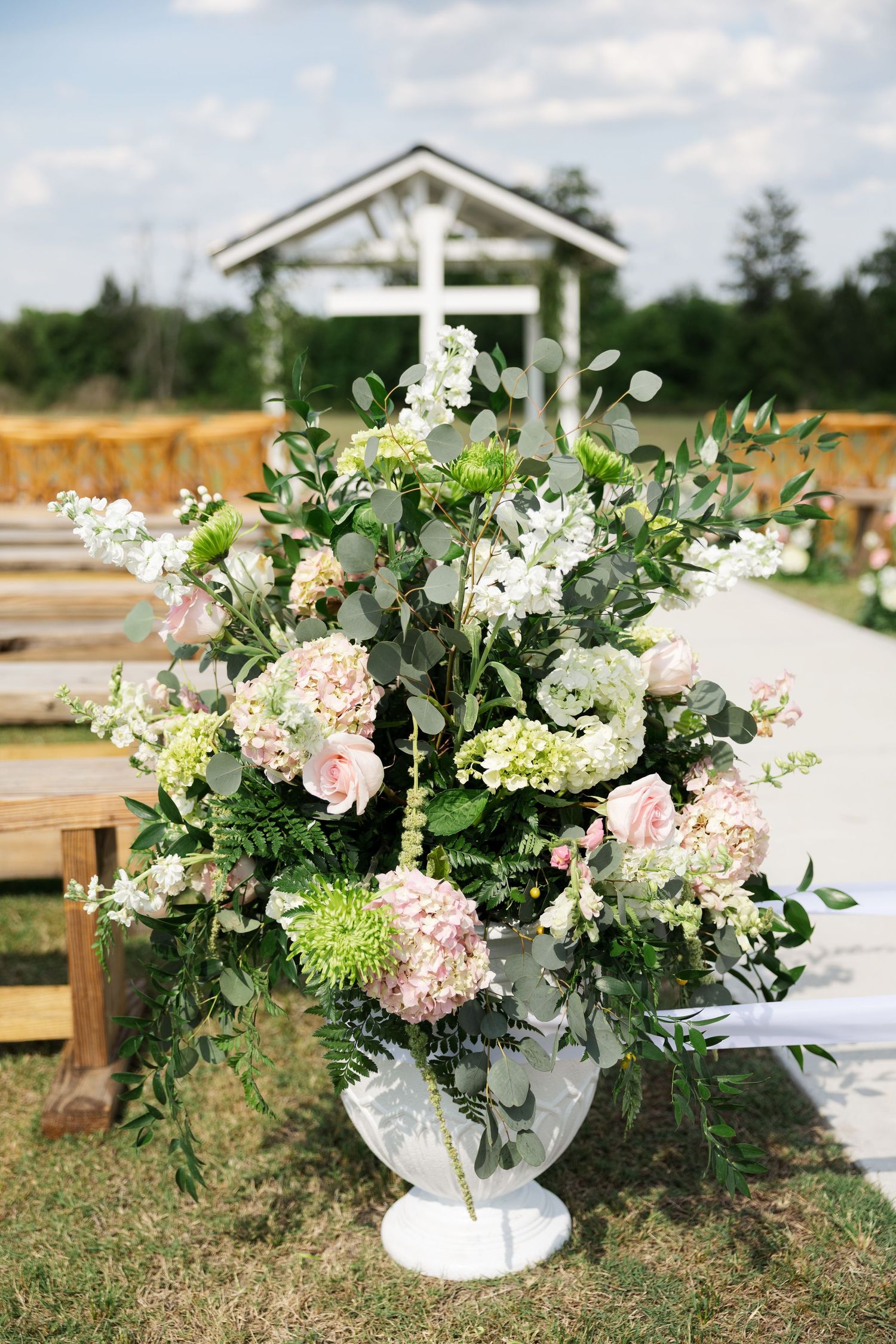 Lush outdoor wedding ceremony aisle arrangement with pink hydrangeas, roses, and greenery in white urn.