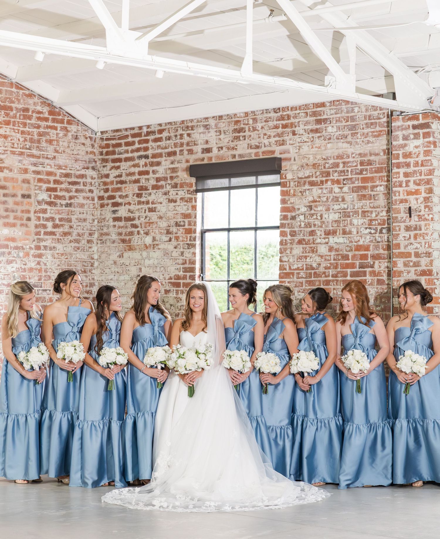 Bride surrounded by bridesmaids in dusty blue satin gowns holding white bouquets in brick venue.