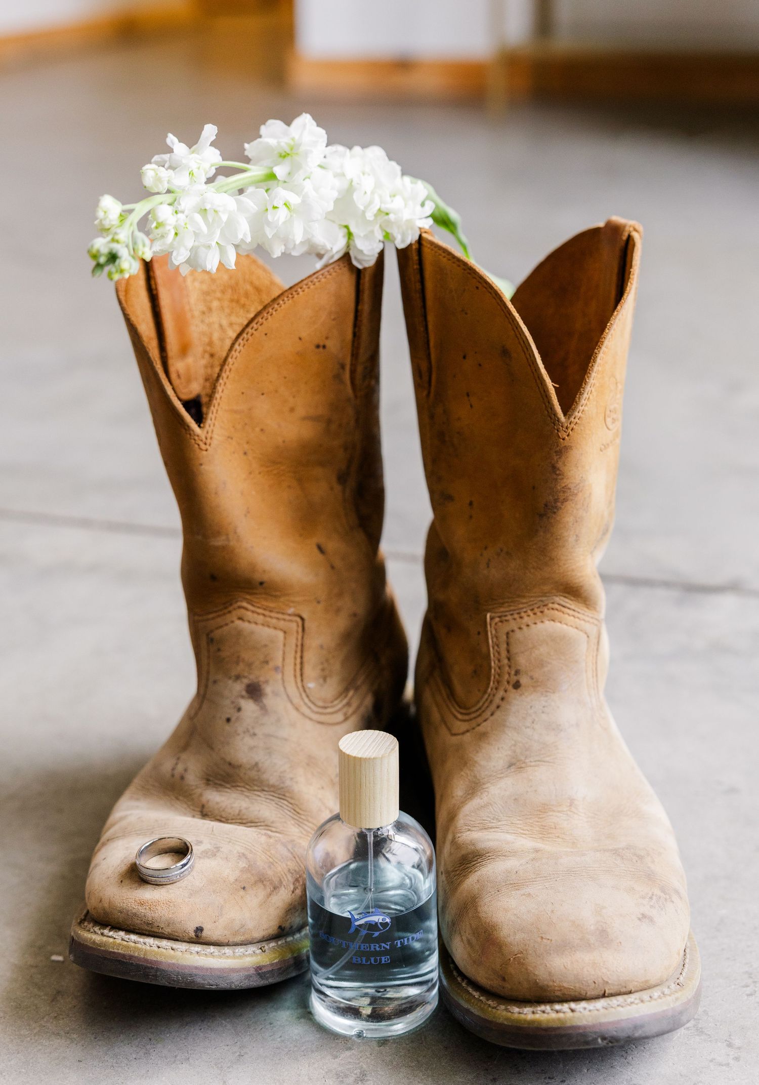 Rustic cowboy boots with white flowers inside, a ring, and a small perfume bottle on a concrete floor.