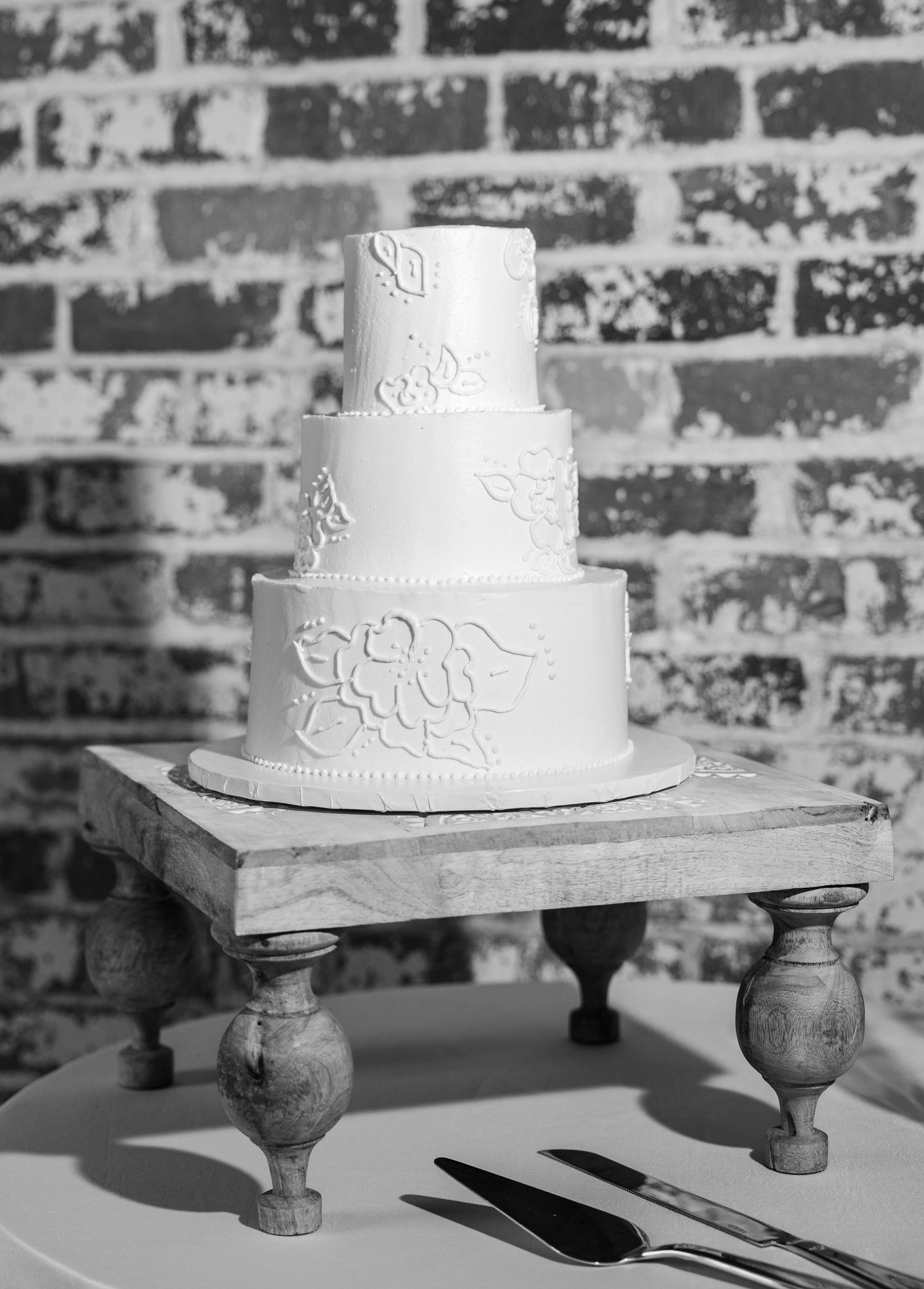 Elegant three-tier white wedding cake with floral details on rustic wood stand against brick wall.