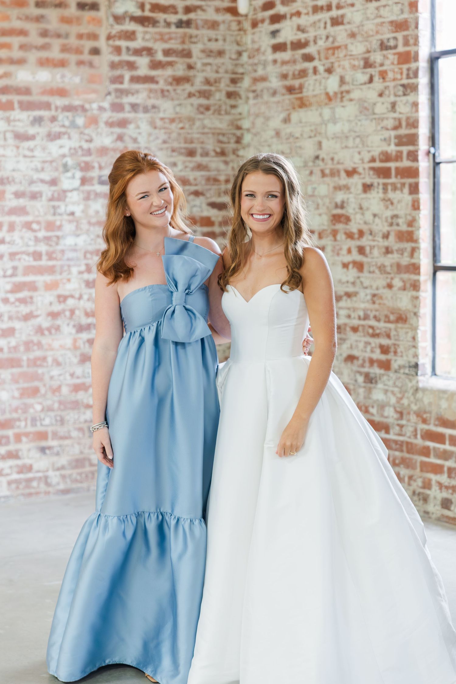 Bride in strapless white ballgown smiles with bridesmaid in dusty blue bow gown against brick wall.