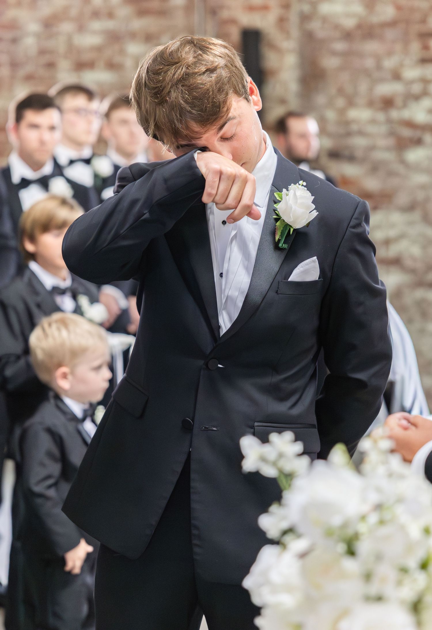 Emotional groom wiping tears at wedding altar wearing black tuxedo with white boutonniere during ceremony.