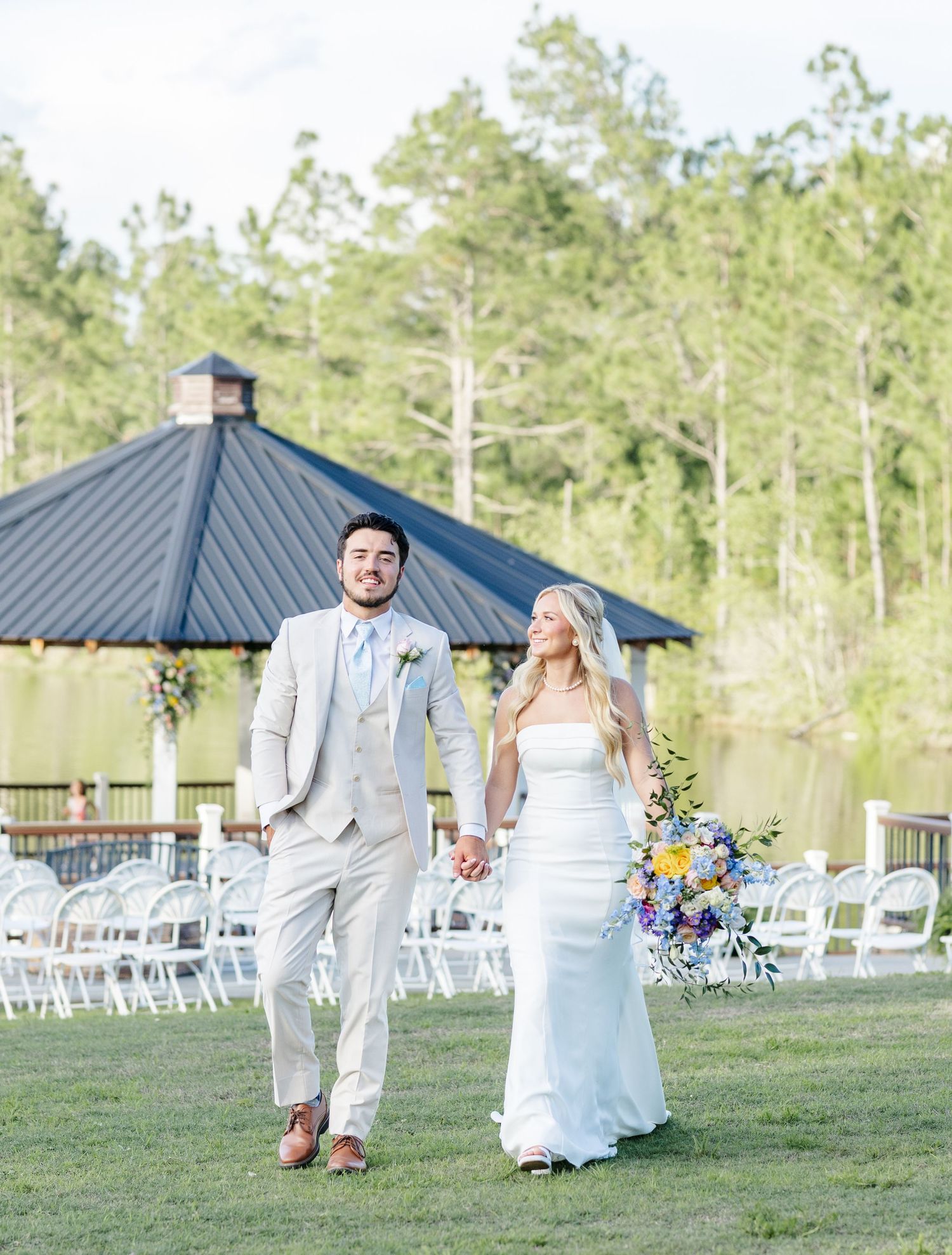 Newlyweds walk hand in hand on green lawn with lakeside pavilion and white chairs in background.