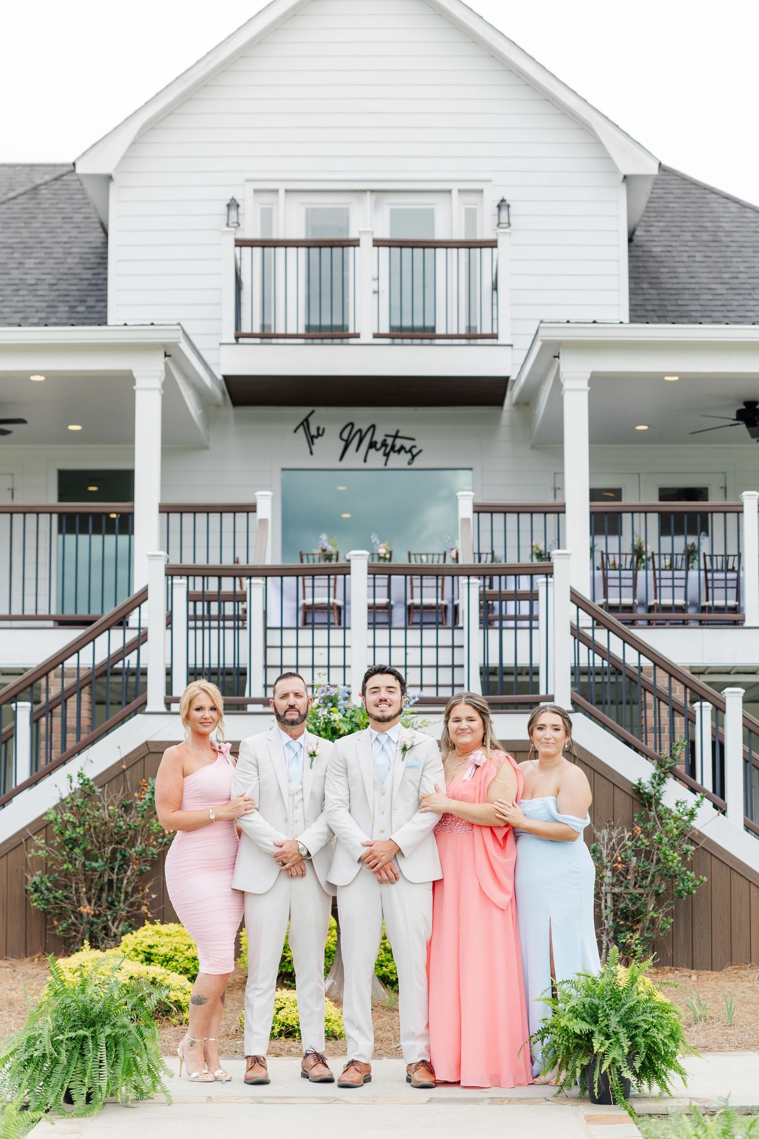 Groom in tan suit poses with wedding party in pink and blue dresses outside white farmhouse venue.