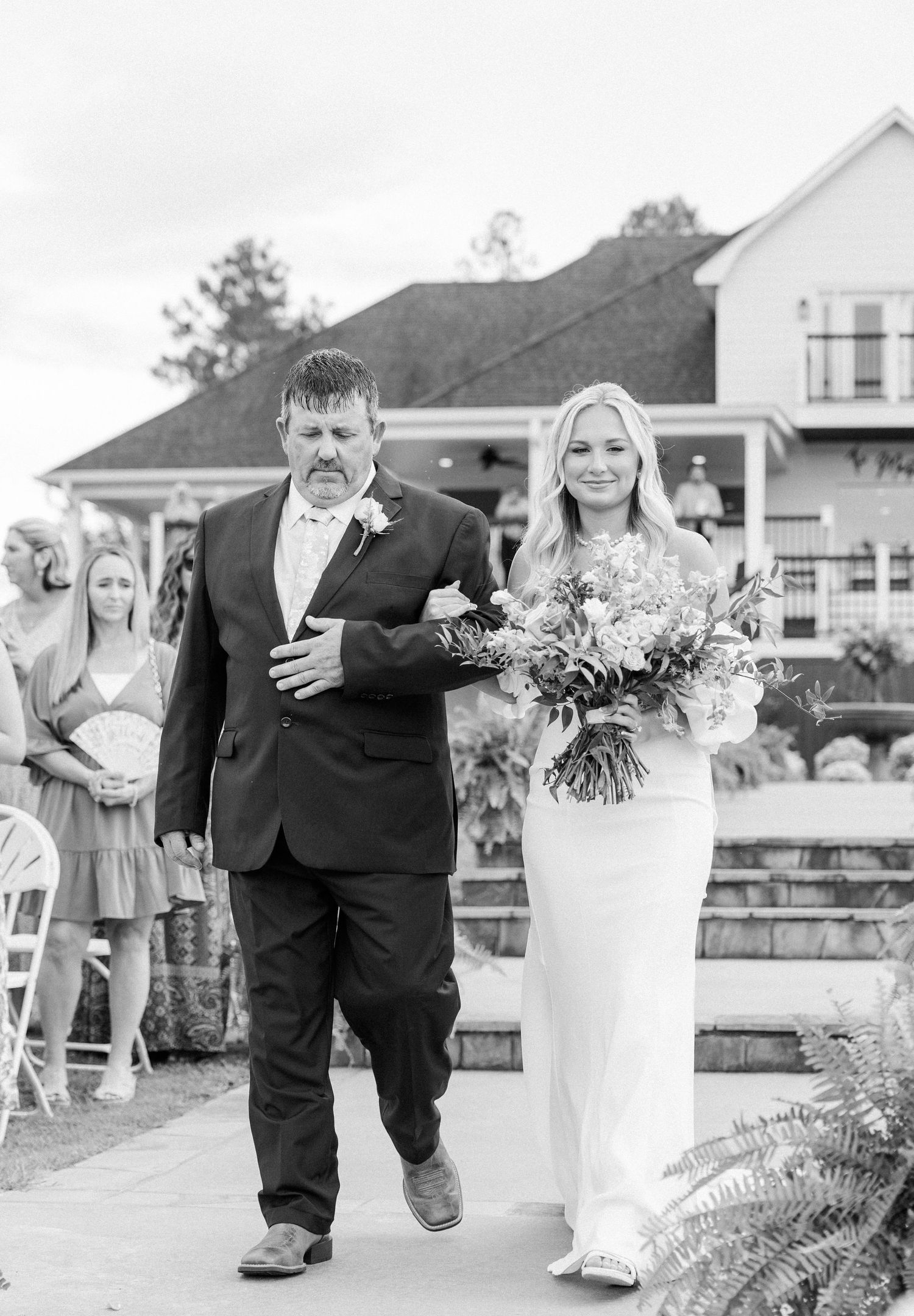 Black and white photo of bride in sleek gown walking down aisle with father holding bouquet.