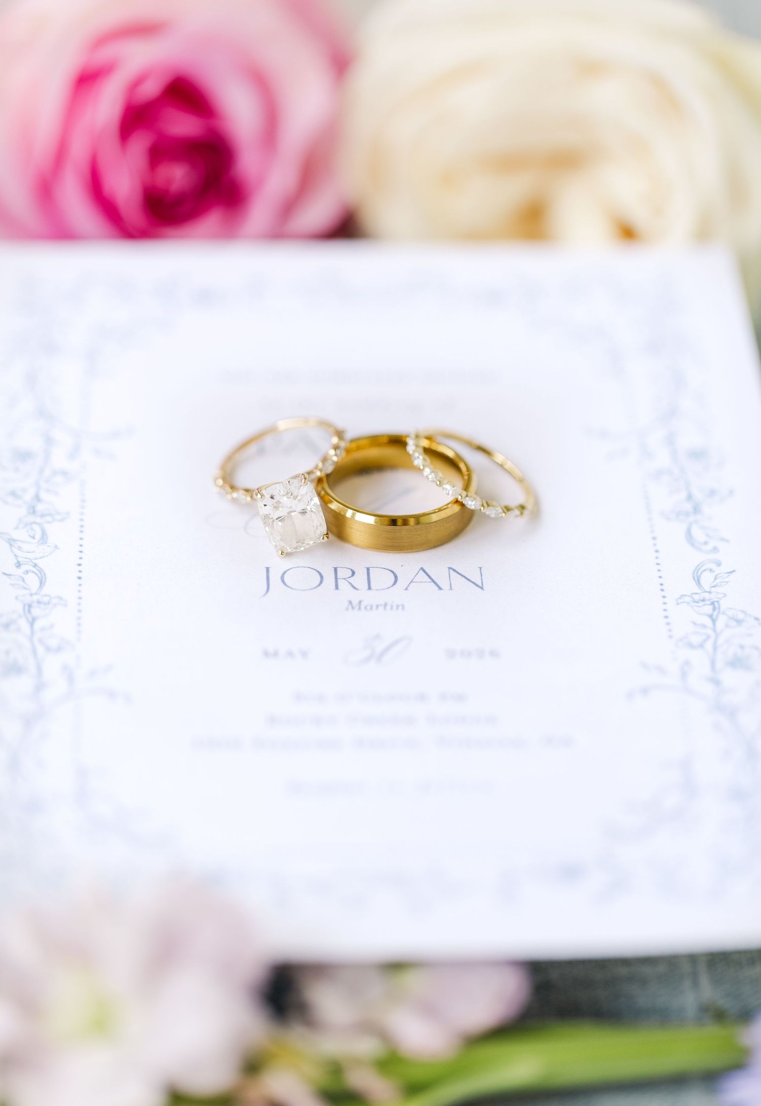 Gold wedding rings and diamond engagement ring resting on elegant wedding invitation with floral accents.