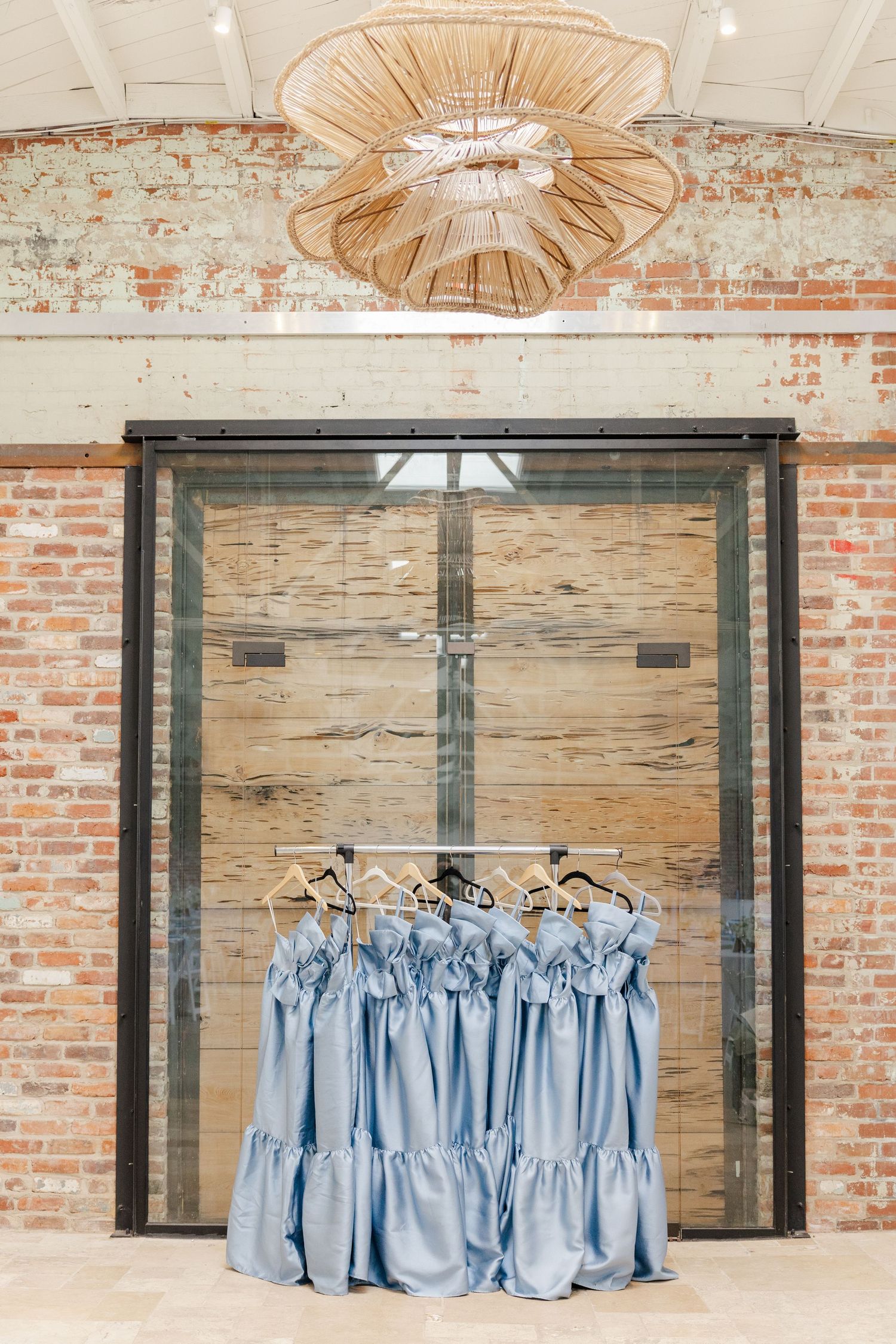 Blue satin bridesmaid dresses hanging on wooden hangers in front of glass doors in a rustic brick venue.