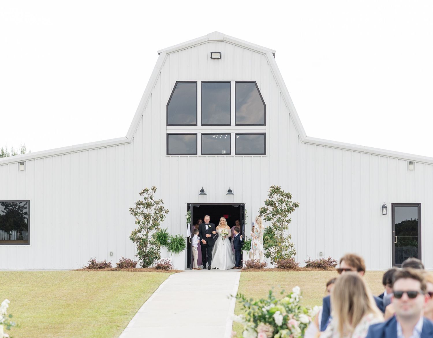 Bride in white gown emerging from white barn wedding venue doors with wedding party for outdoor ceremony.
