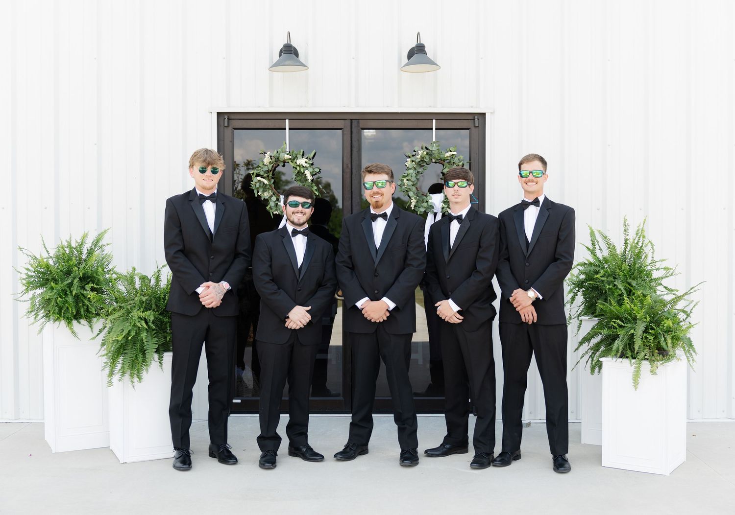 Five groomsmen in black tuxedos and masquerade masks pose together outside a white barn wedding venue.