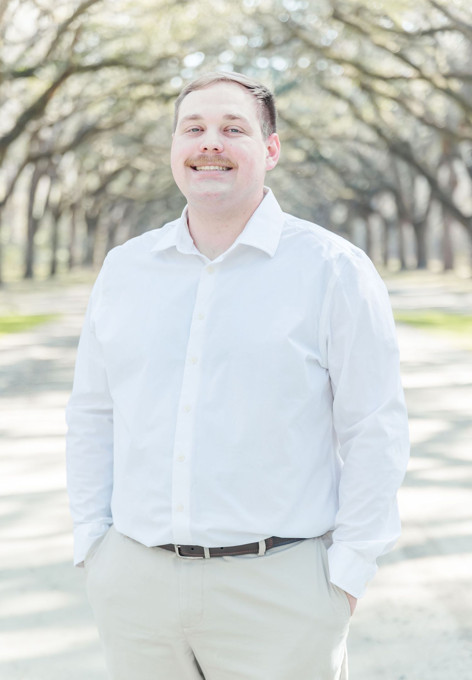 A man in white dress shirt and khakis poses on a tree-lined path.