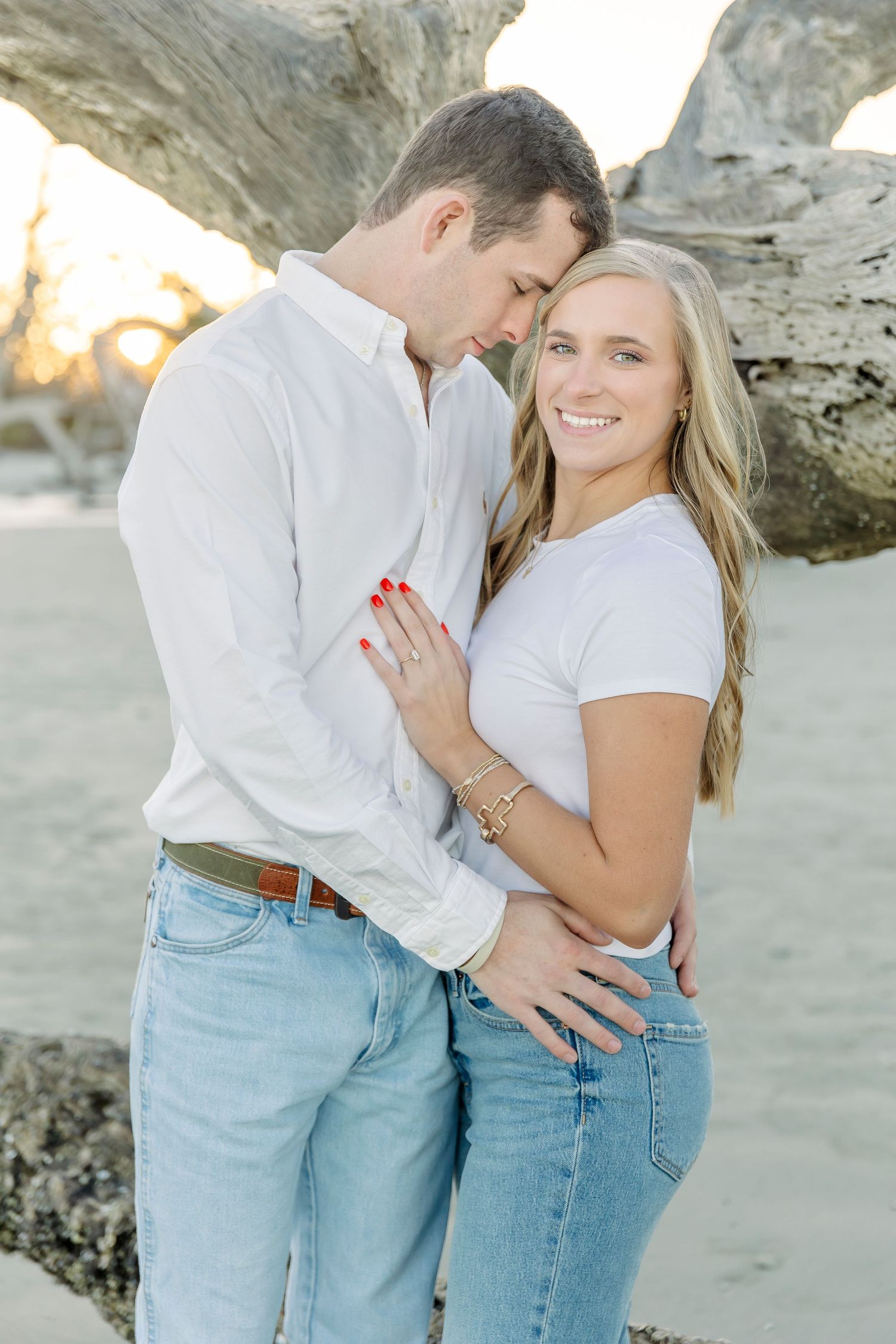 A couple in white and denim shares an intimate moment against driftwood on the beach.