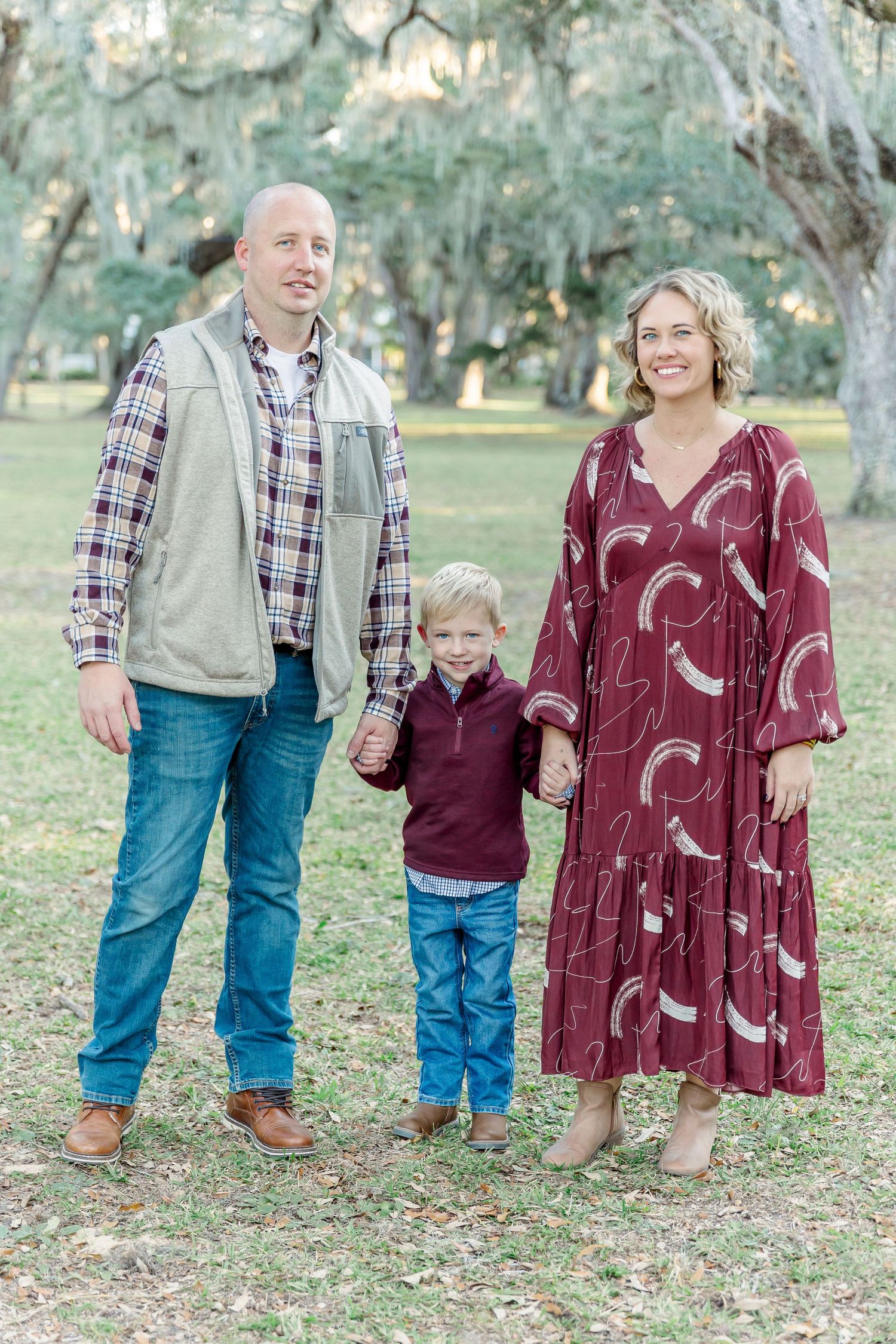 Family of three poses in casual attire under Spanish moss covered trees in a park setting.