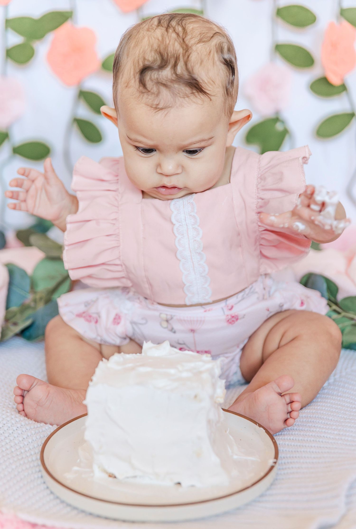 Baby in pink ruffled outfit reaching toward small white cake on plate.