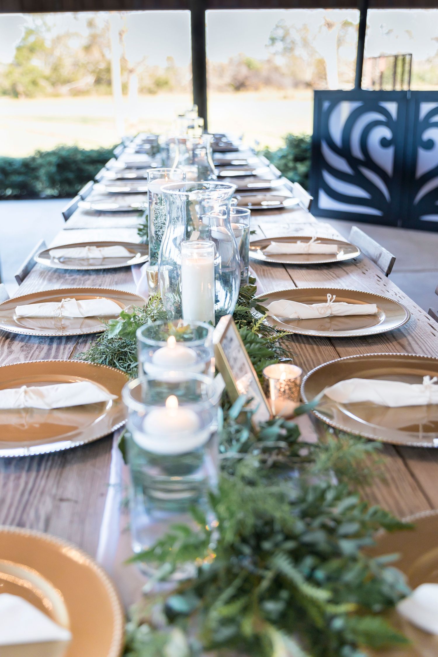 Long dining table with gold plates and glass candle holders decorated with fresh greenery garland.