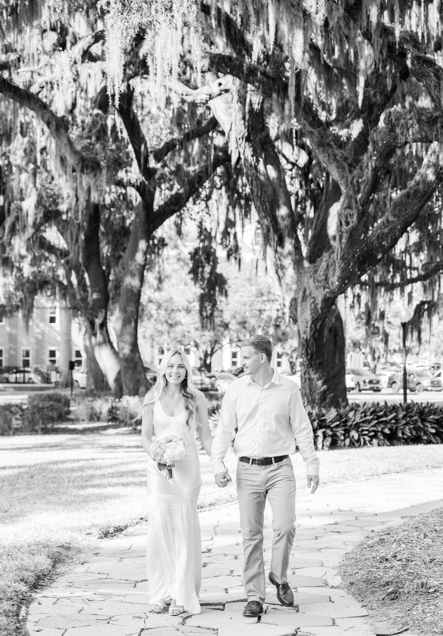 Black and white photo of a couple walking together on a stone path under moss-covered trees.
