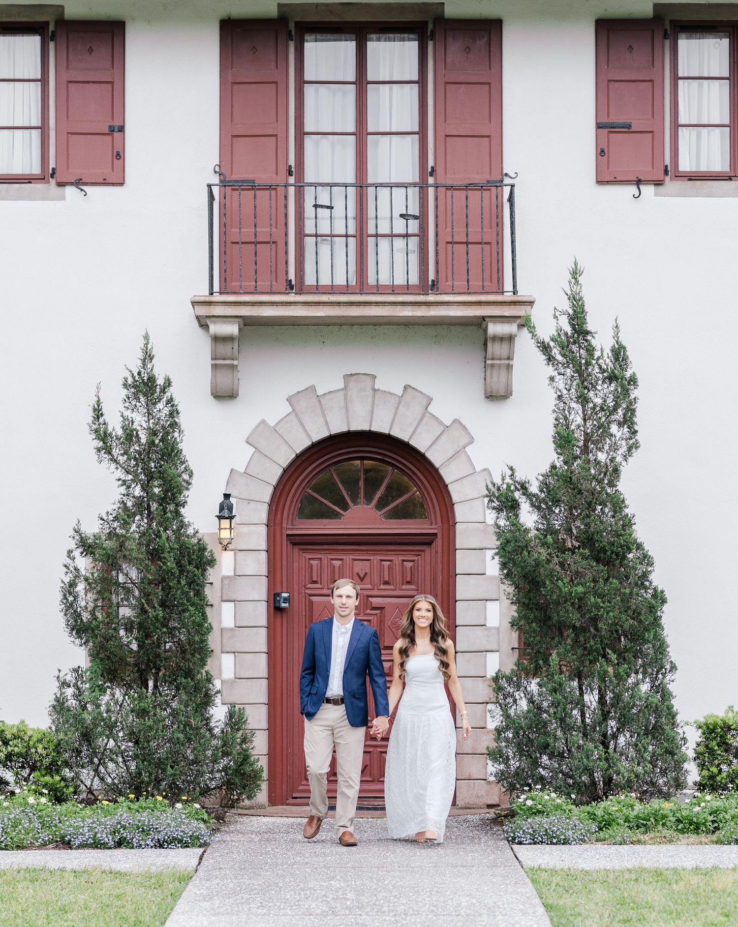 Couple holding hands in front of elegant home with red arched door, tall cypress trees, and balcony above.