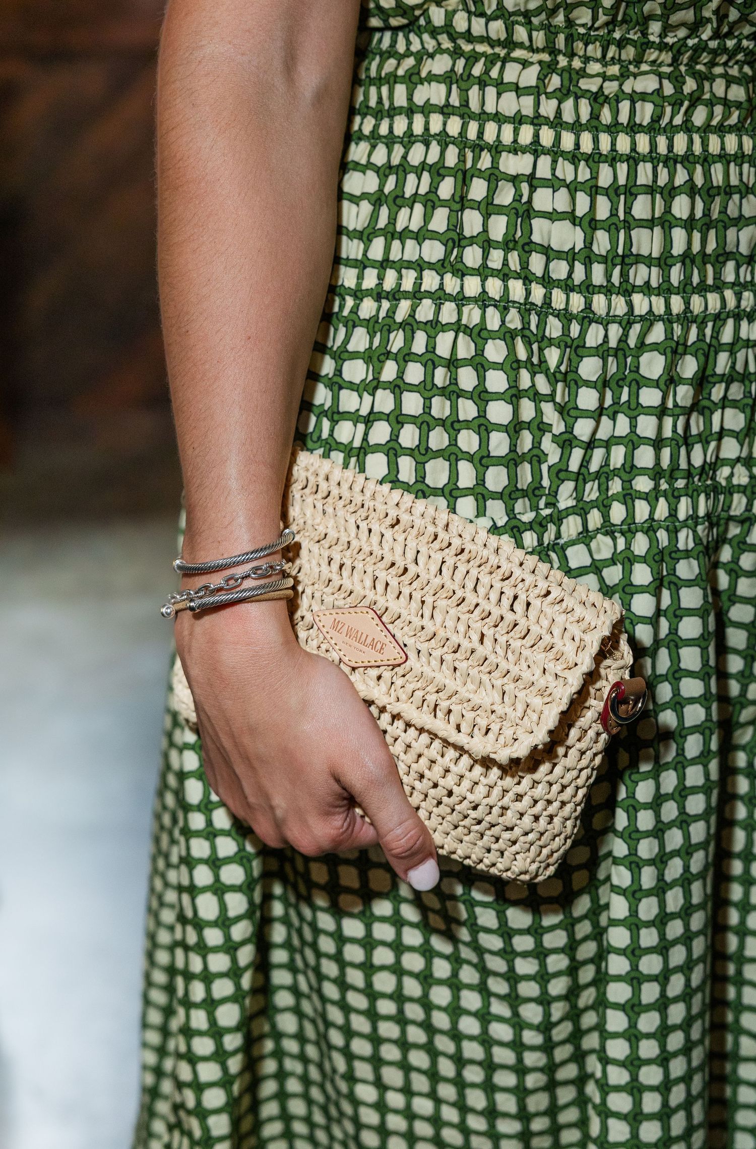 Woman in green printed dress holding a woven raffia clutch bag with silver bracelet accessory.