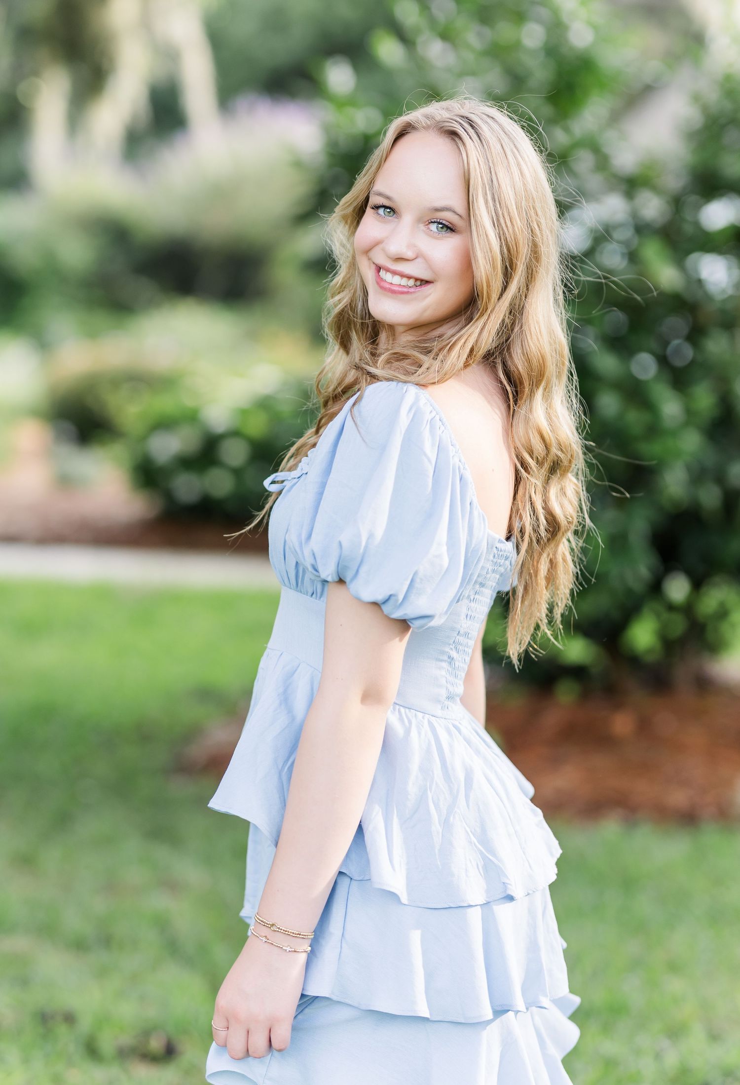 A person in a light blue ruffle dress poses in a garden with a bright smile and flowing hair.
