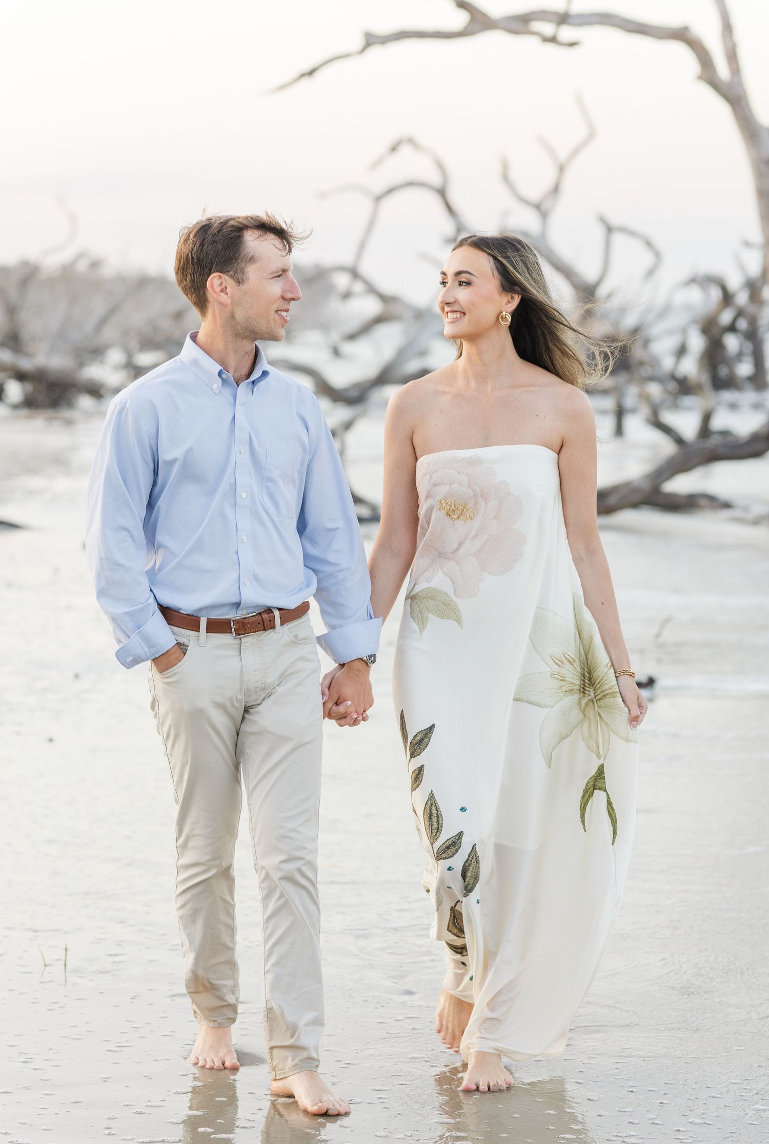 Couple holding hands walking barefoot on beach, woman in white floral strapless gown, man in light blue shirt.