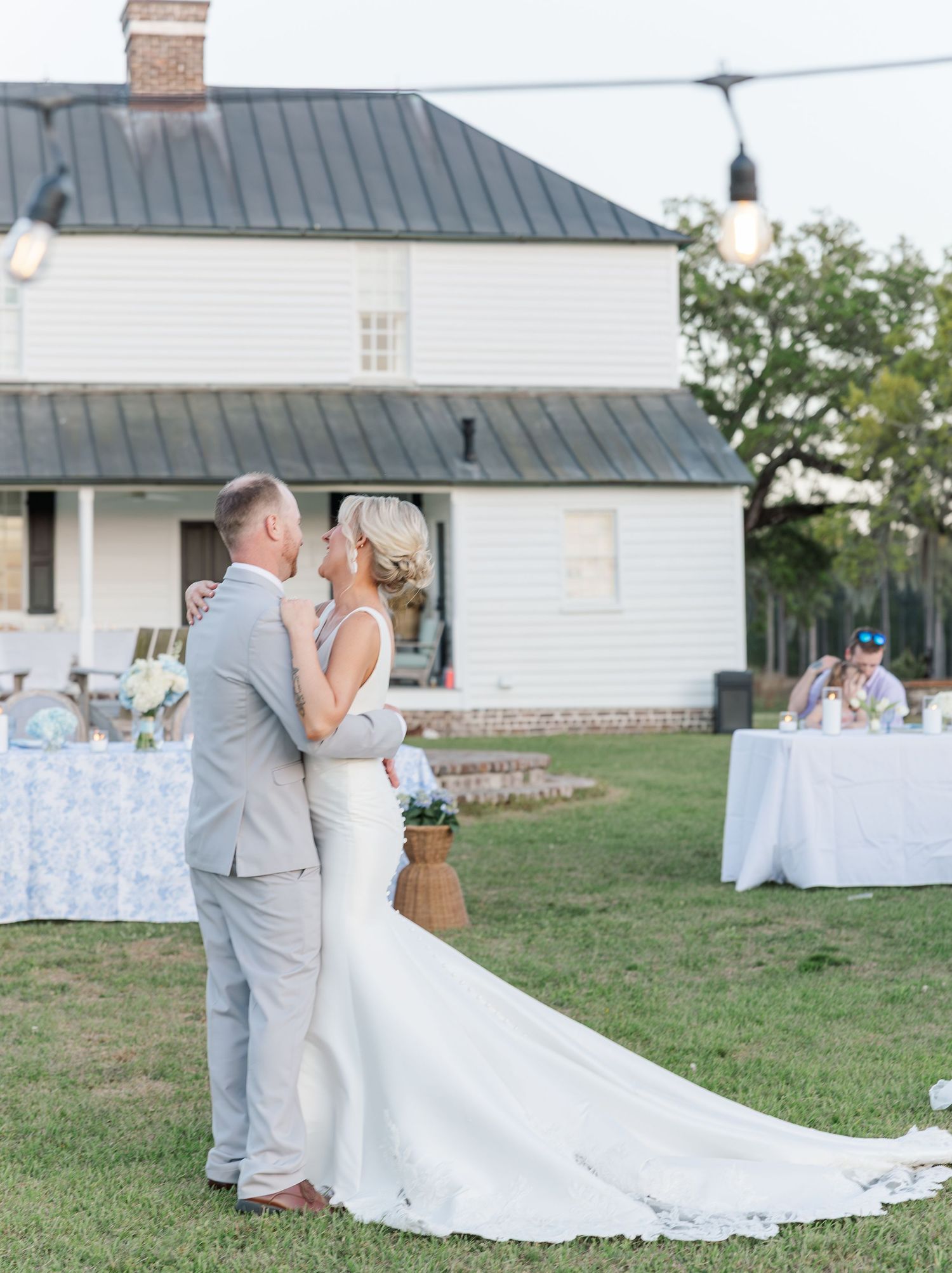 Bride and groom share first dance on lawn at dusk in front of white farmhouse with string lights.