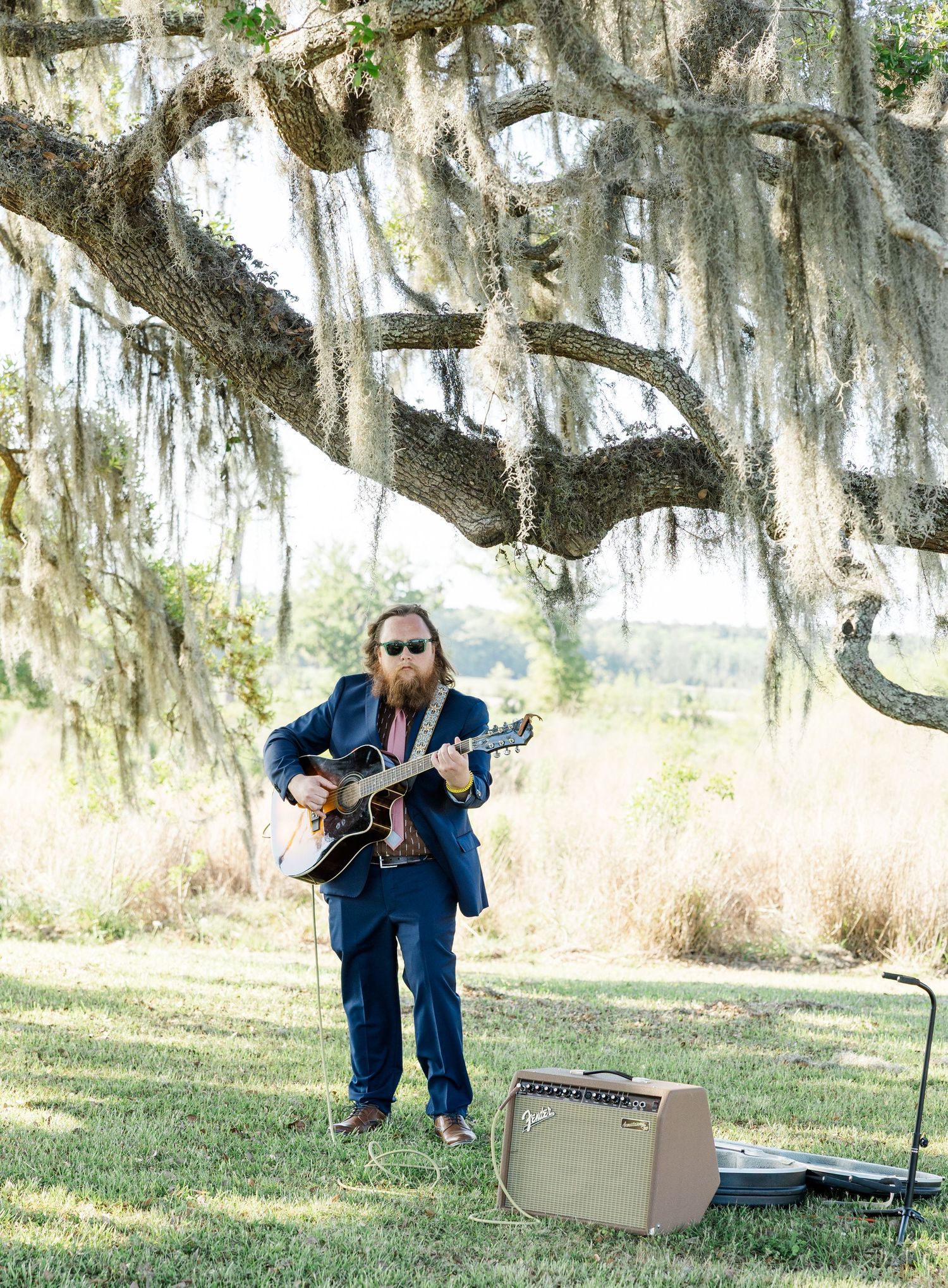 Bearded musician in navy suit plays guitar under Spanish moss-draped oak tree at outdoor wedding ceremony.