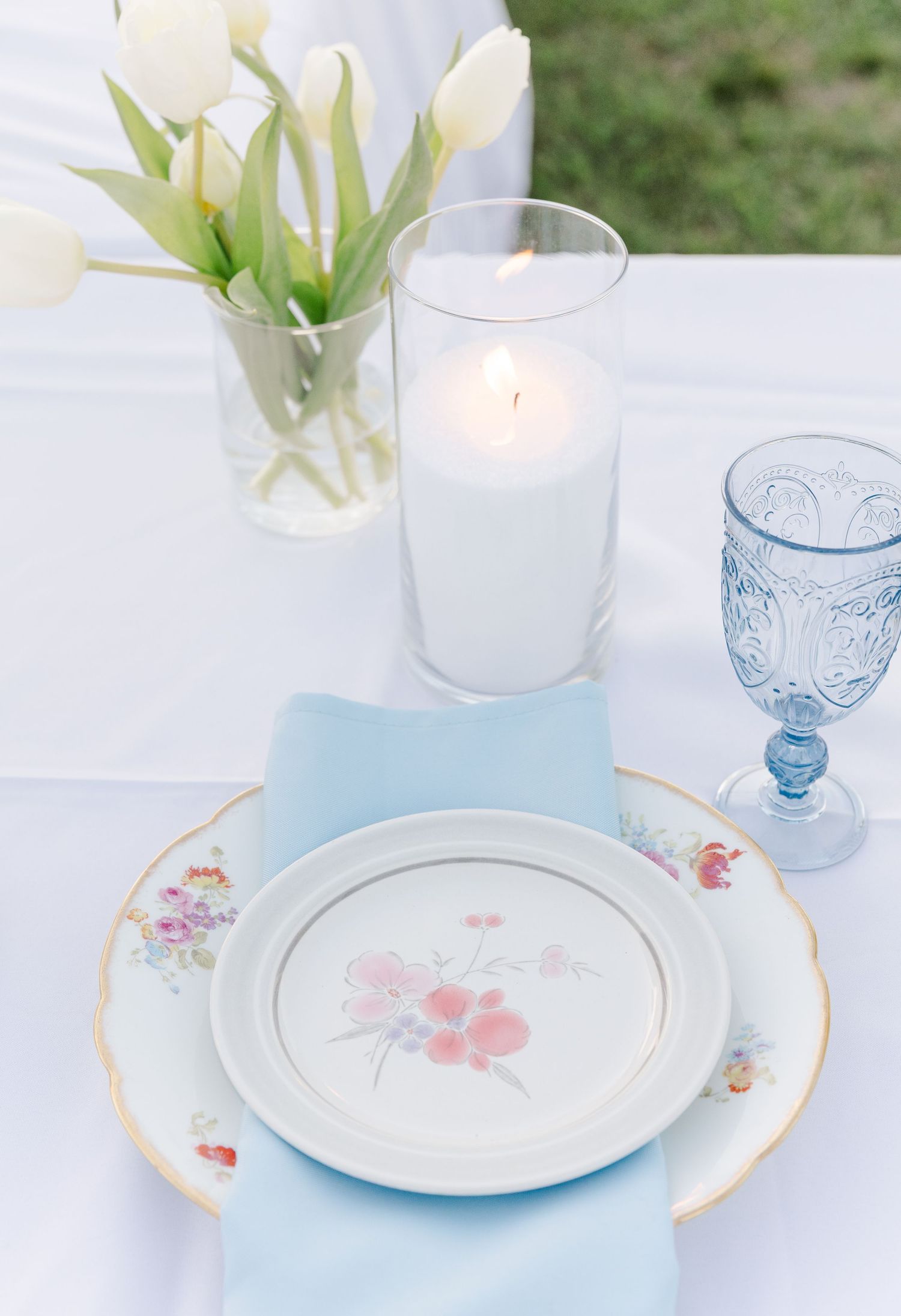 Elegant wedding table setting with floral plates, blue napkin, lit candle, white tulips, and blue goblet.