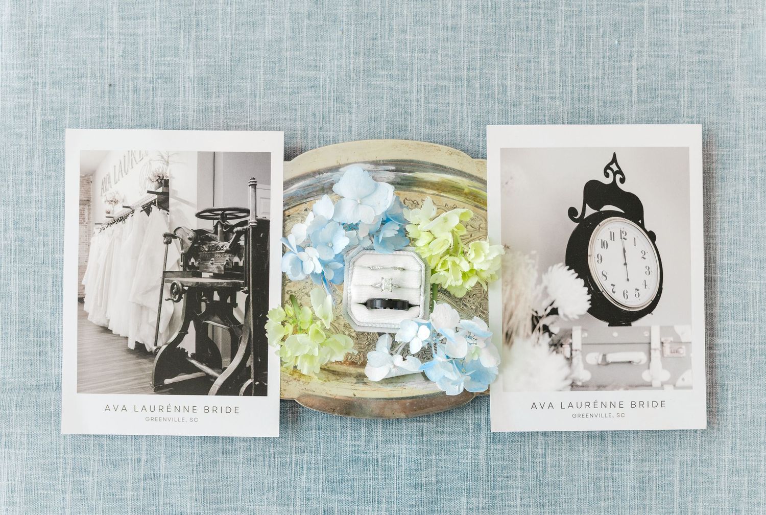 Wedding flat lay featuring Ava Laurenne Bride photos, blue hydrangeas, green florals, ring, and vintage clock on blue linen.