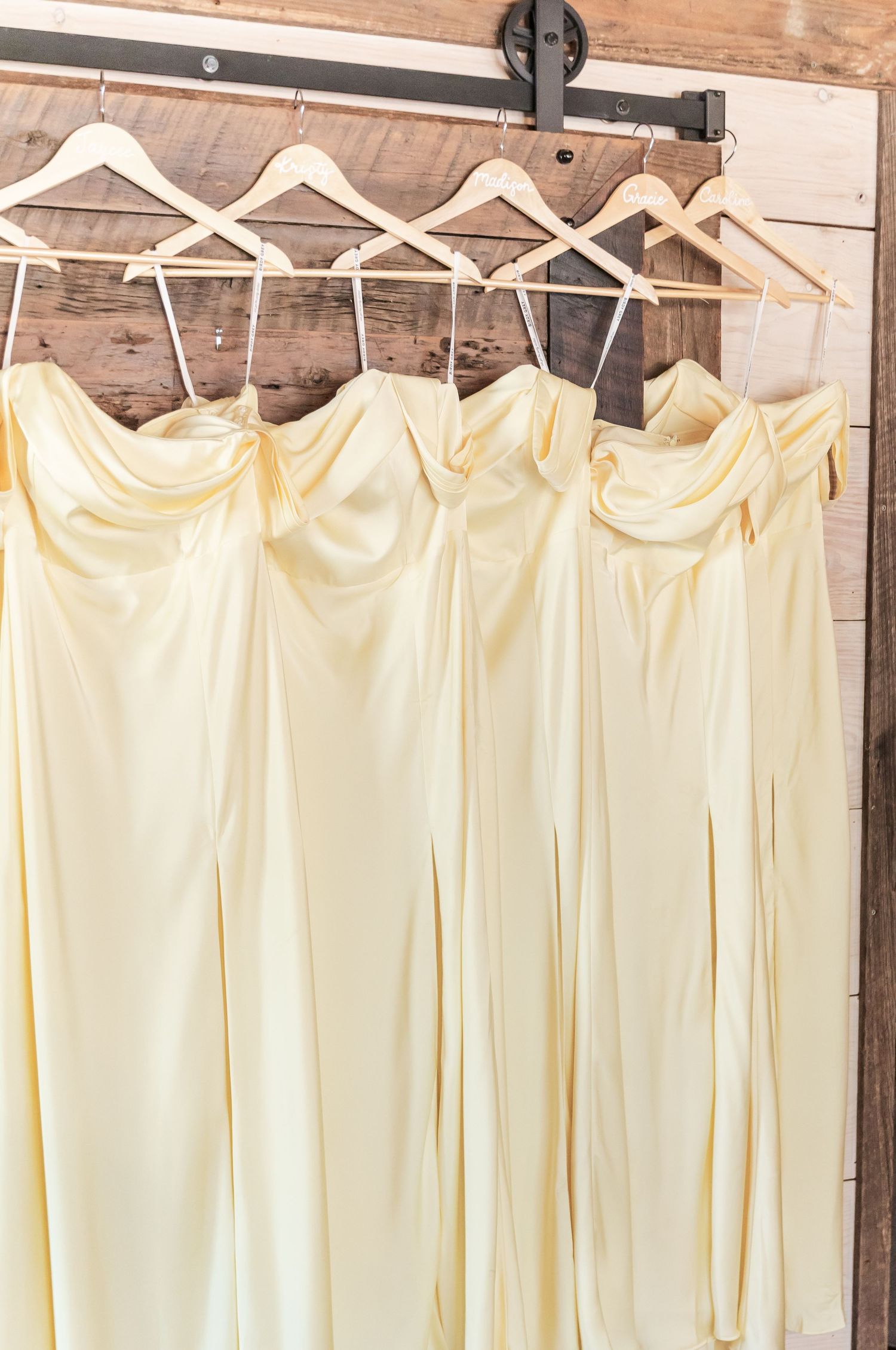 Five yellow strapless bridesmaid dresses hanging on wooden hangers against a rustic barn wall.