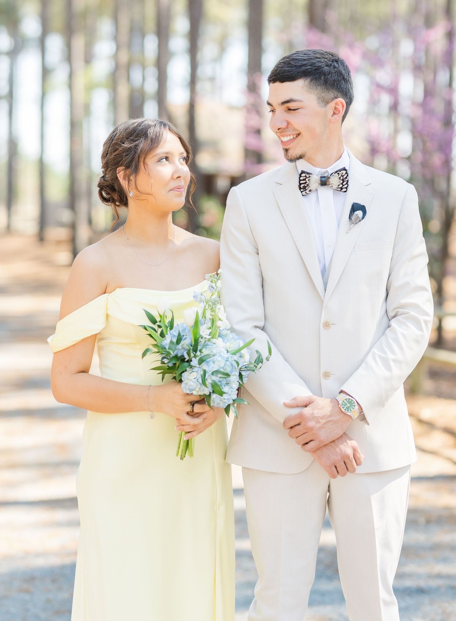 Bridesmaid in yellow off-shoulder gown with groomsman in beige suit and floral bow tie outdoors.