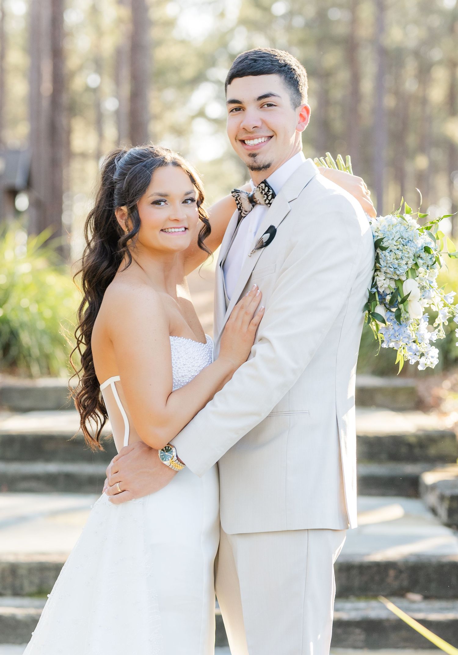 Smiling bride in strapless gown and groom in tan suit holding blue hydrangea bouquet outdoors.