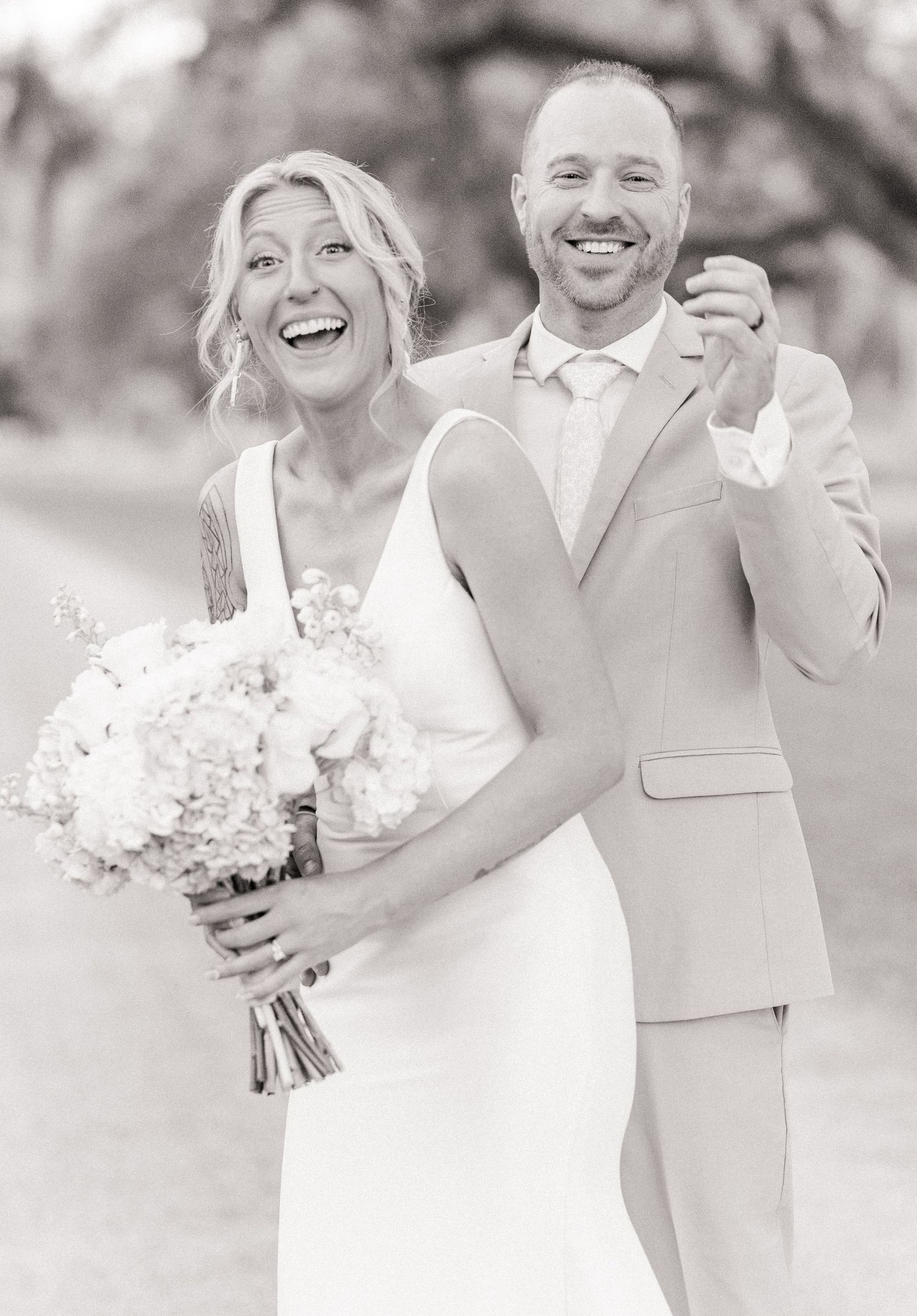Black and white photo of laughing bride and groom celebrating joyfully outdoors with bouquet in hand.