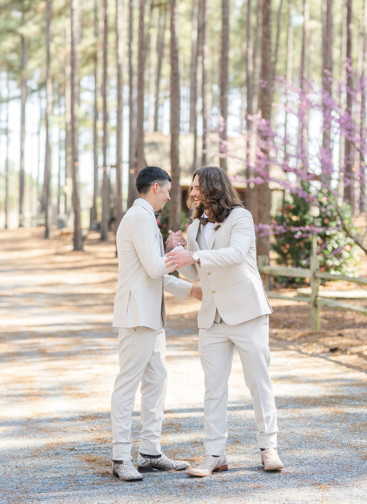 Two groomsmen in ivory suits share a joyful moment on a gravel path amid blooming trees.
