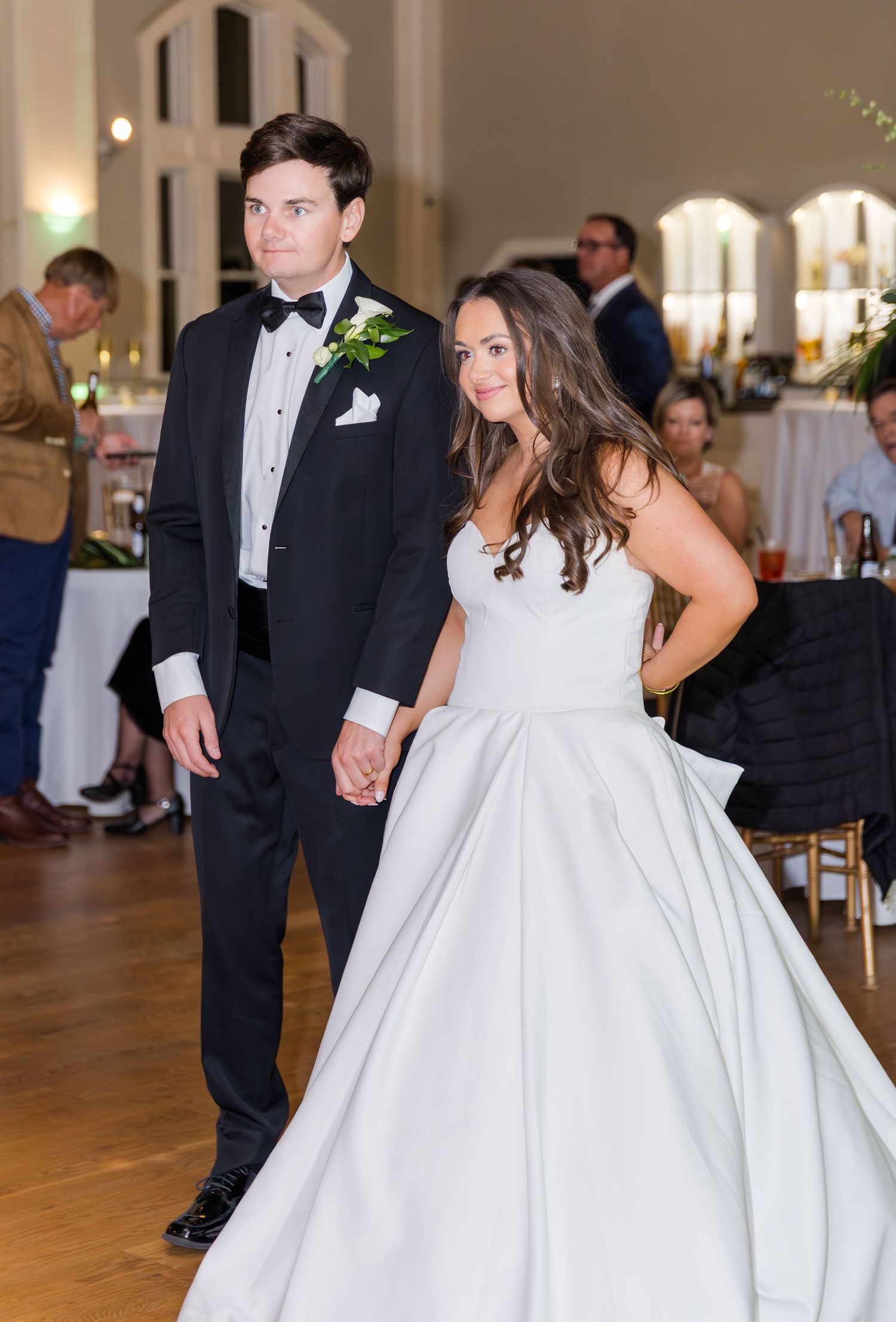 Bride and groom holding hands on dance floor, wearing white ball gown and black tuxedo with floral boutonniere.