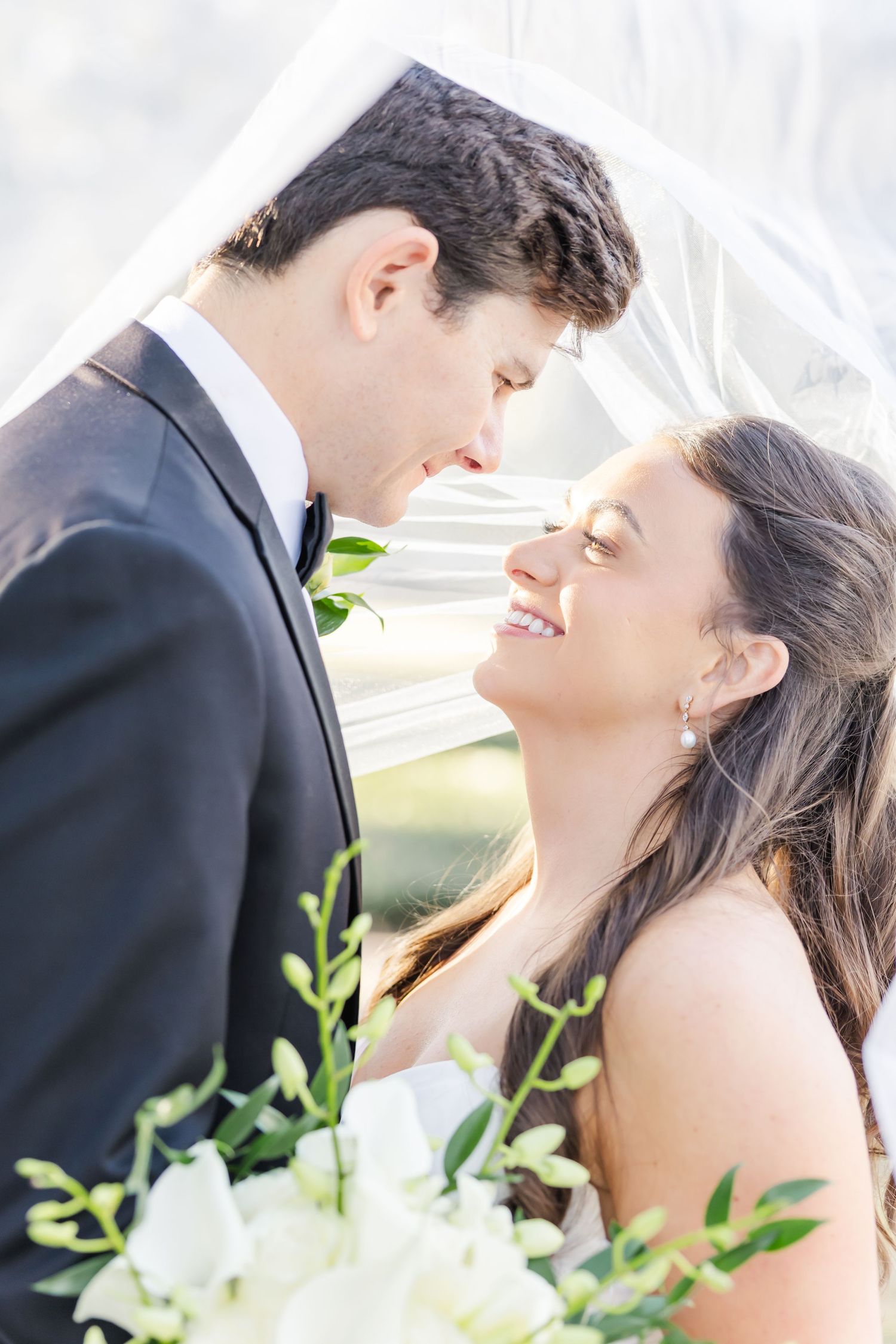 Smiling bride and groom share an intimate moment under a flowing veil with white floral bouquet in foreground.