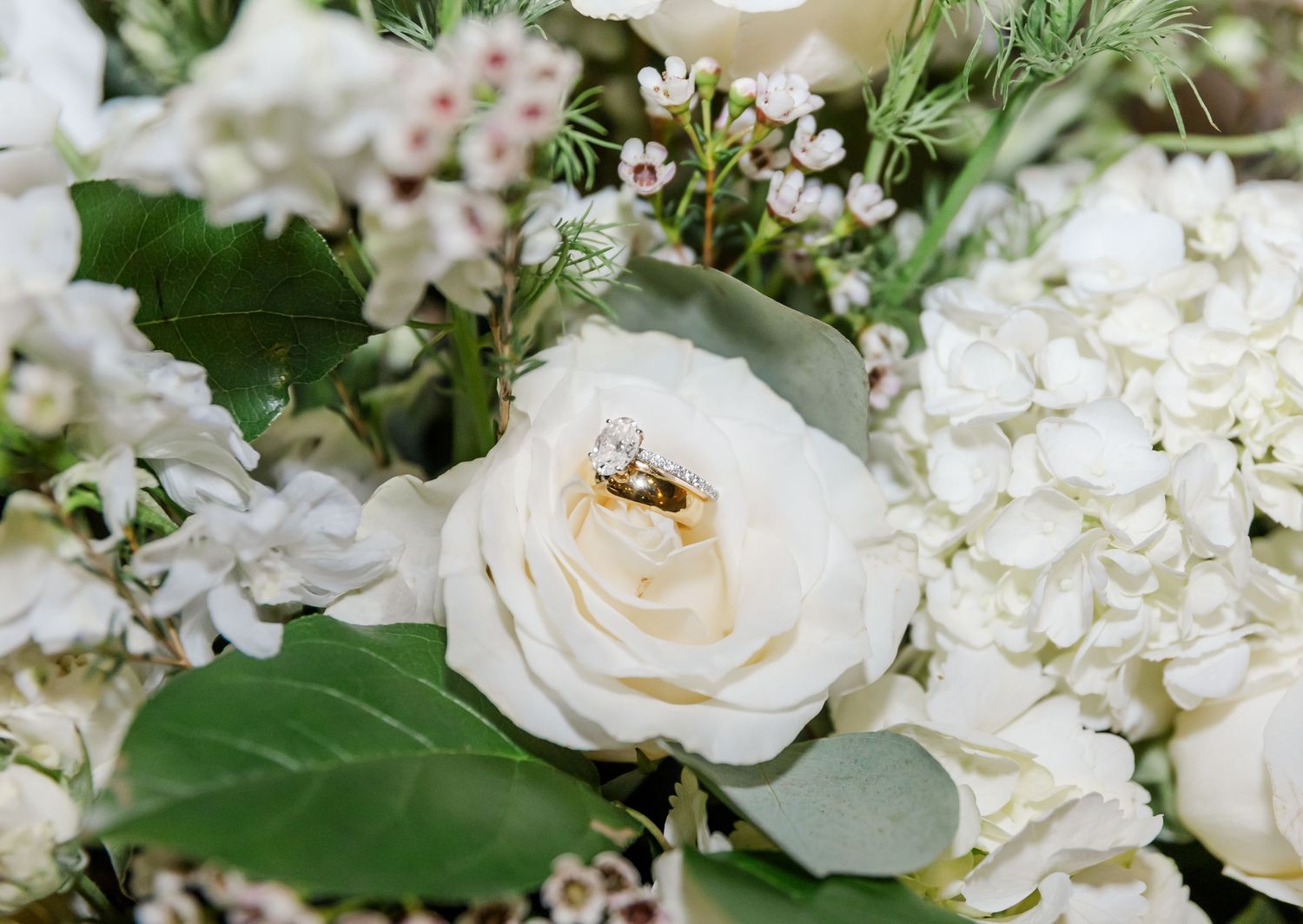Wedding rings nestled in a white rose surrounded by hydrangeas and greenery in a bridal bouquet.