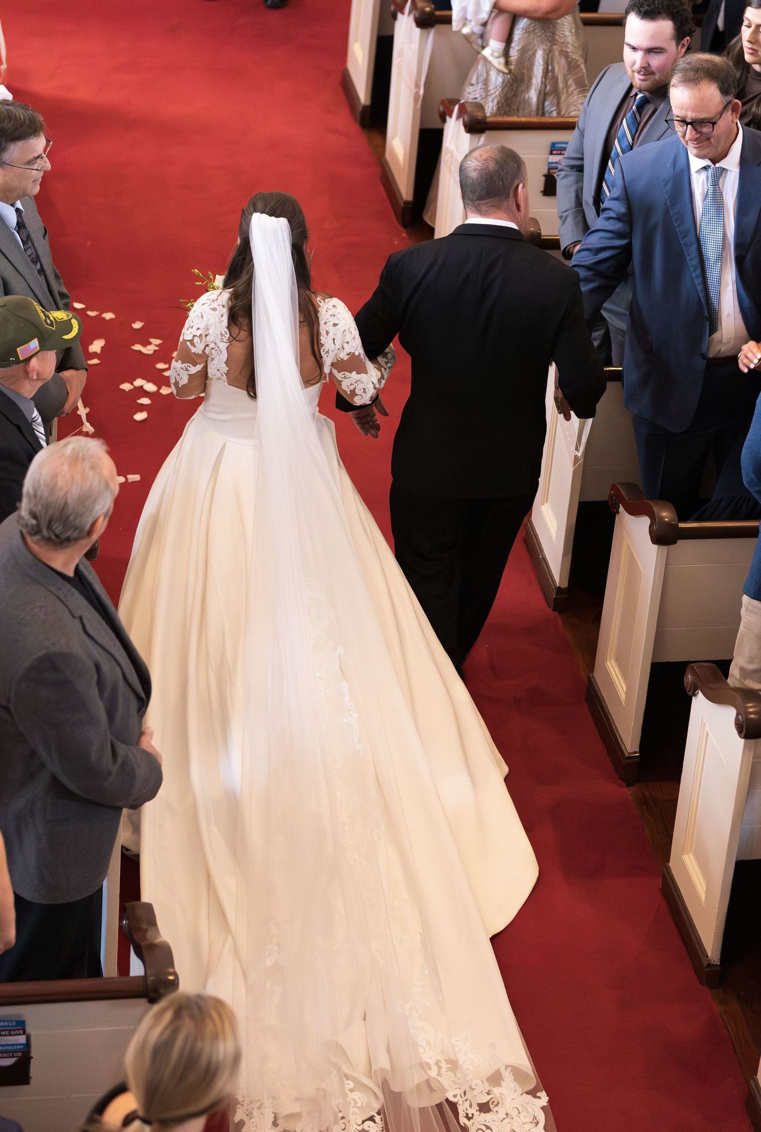 Bride in white gown and veil walks down church aisle with escort during wedding ceremony.