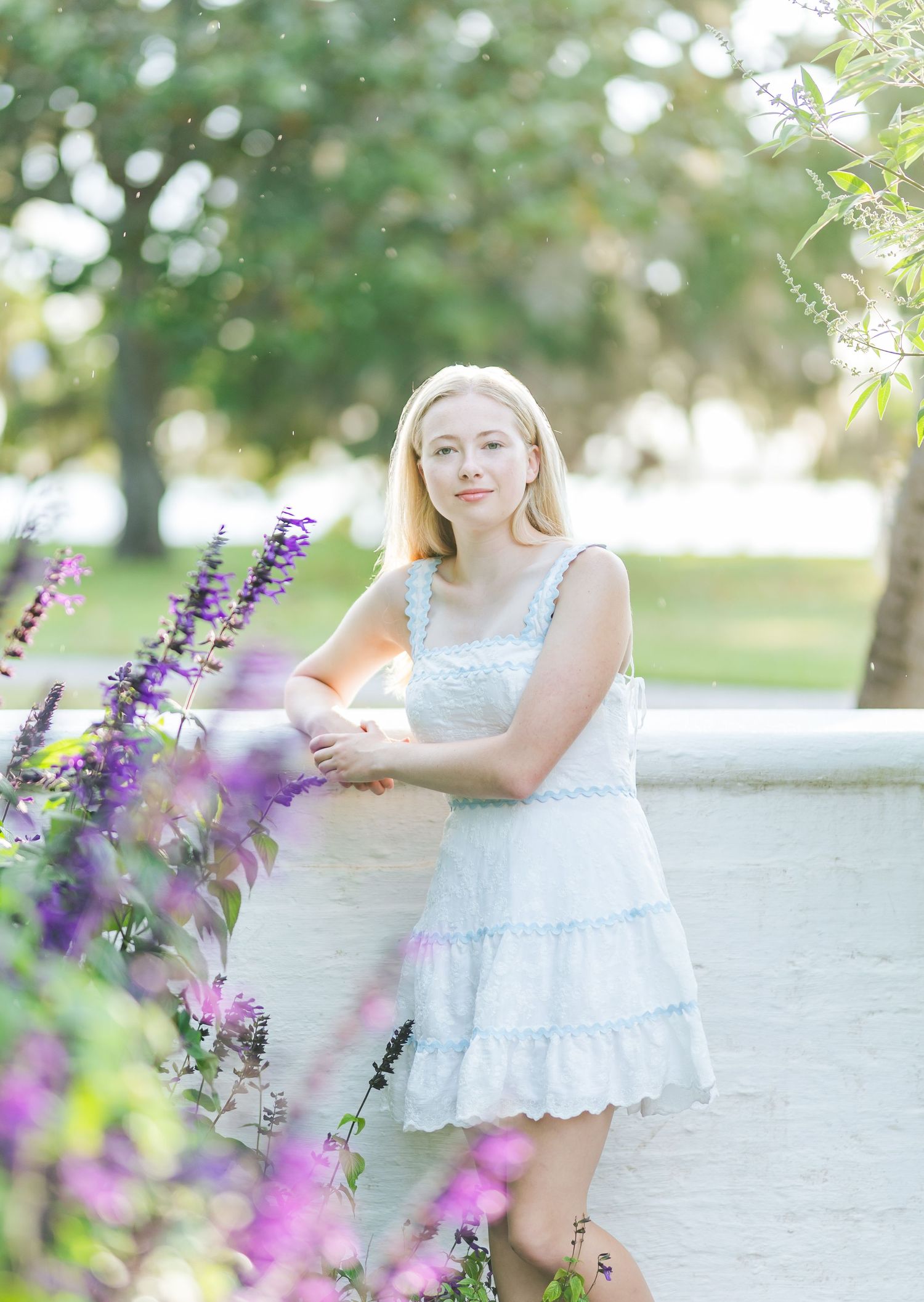 A child in a white lace dress plays near blooming purple salvia flowers in a sunny garden setting.