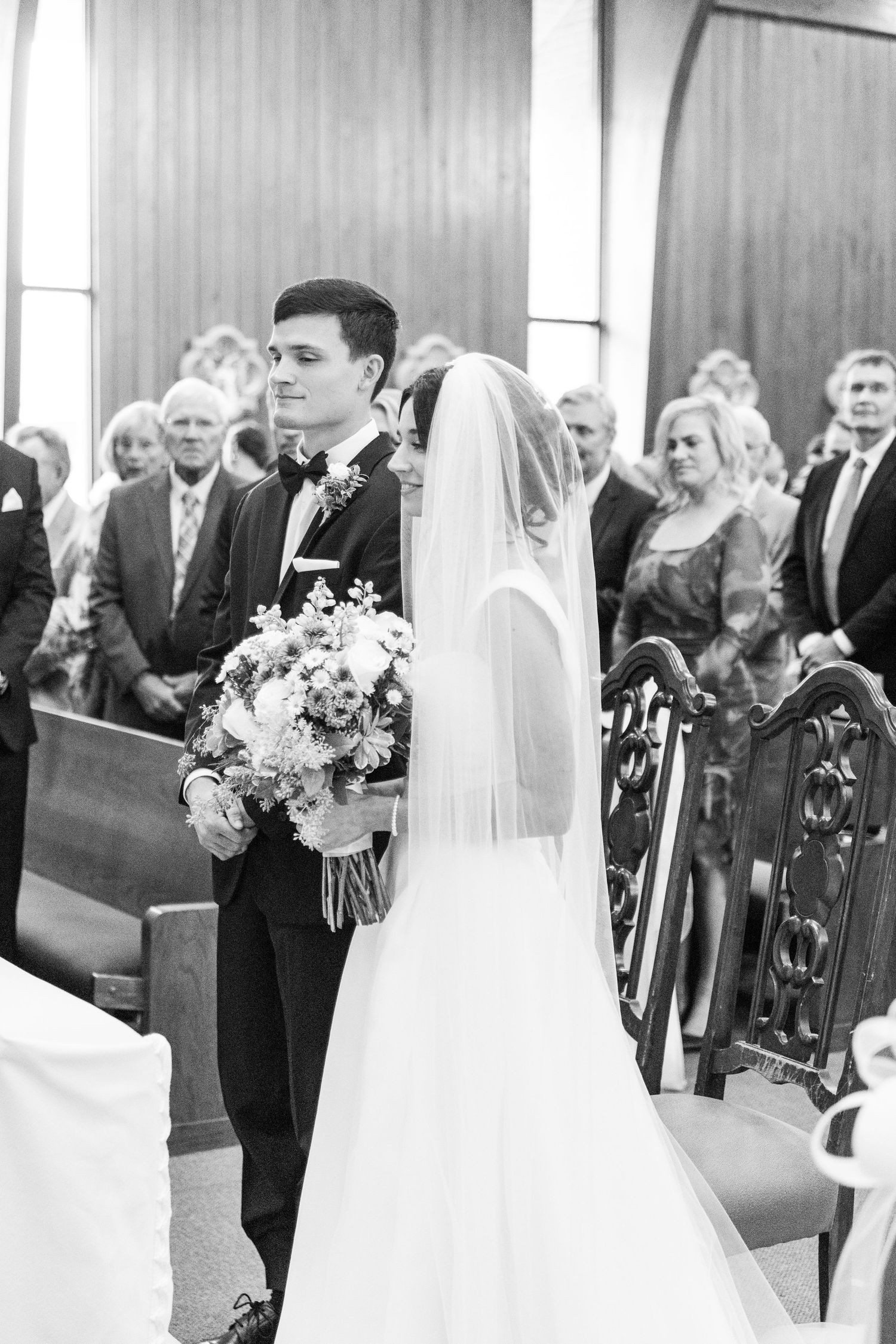 Church wedding ceremony with guests seated in ornate wooden pews and floral decorations.