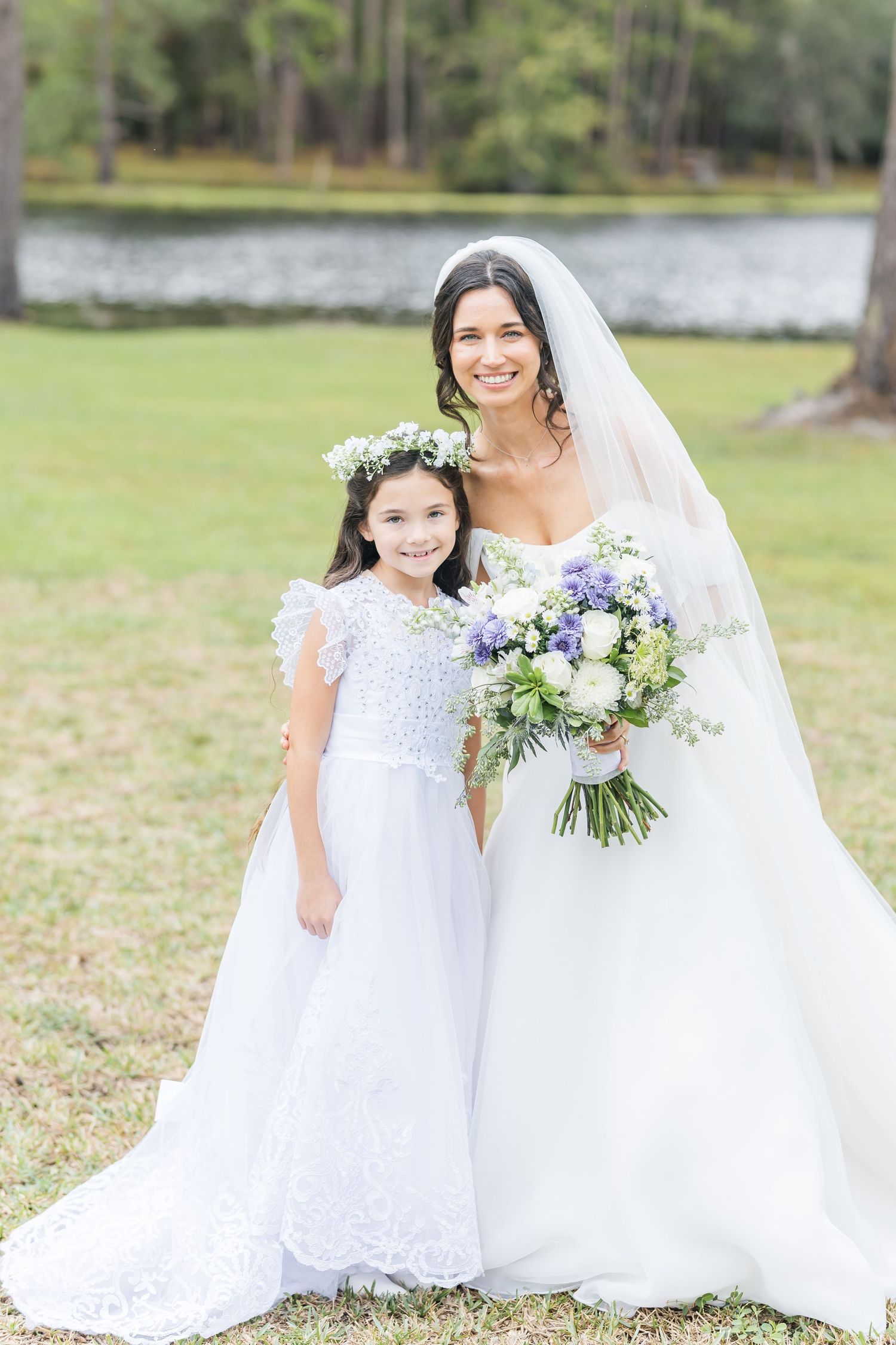 A bride in a white gown poses with a flower girl by a lake holding a beautiful purple and white bouquet.
