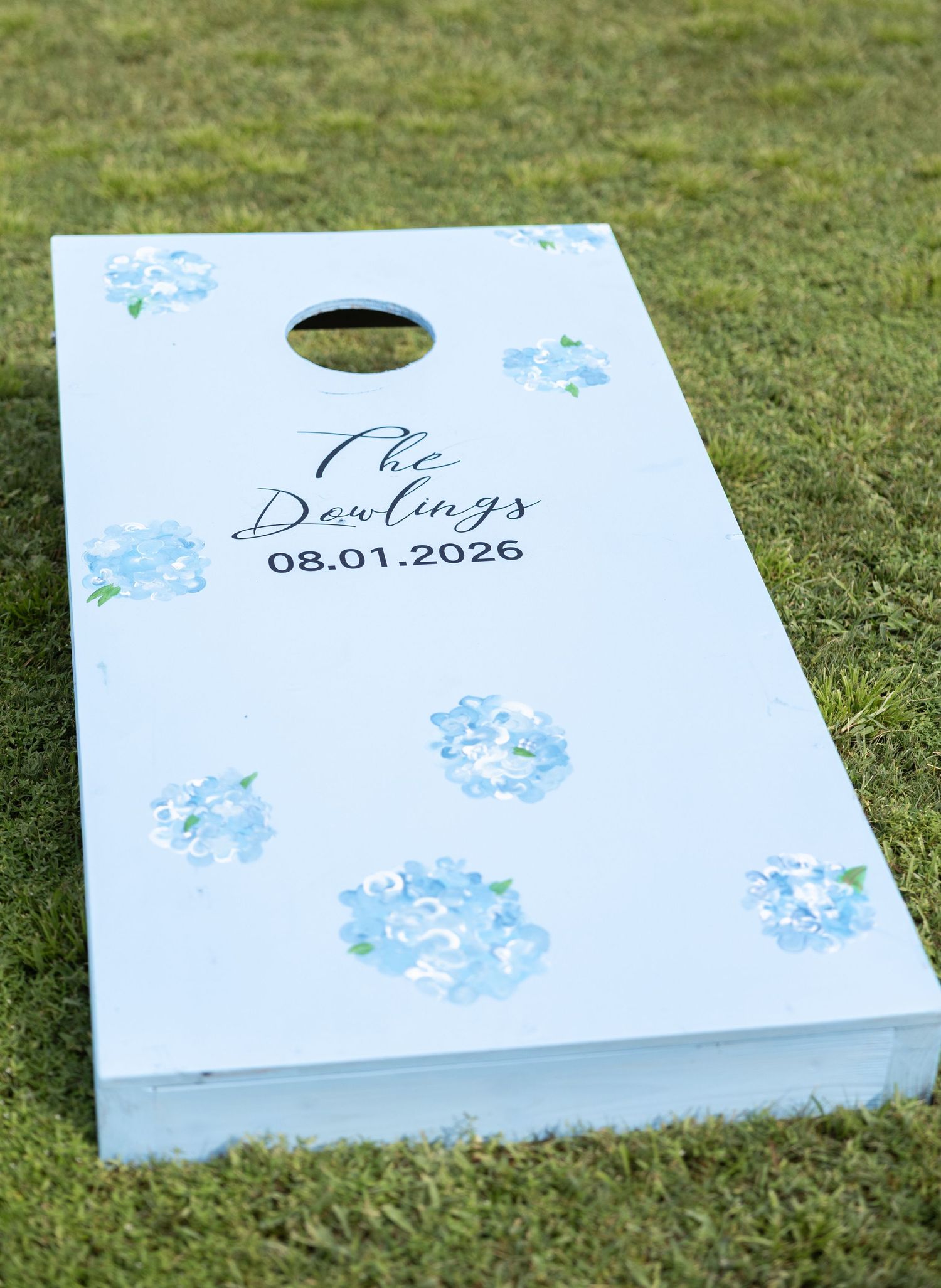 Custom white cornhole board with blue hydrangea florals reading The Dowlings 08.01.2026 on green grass.