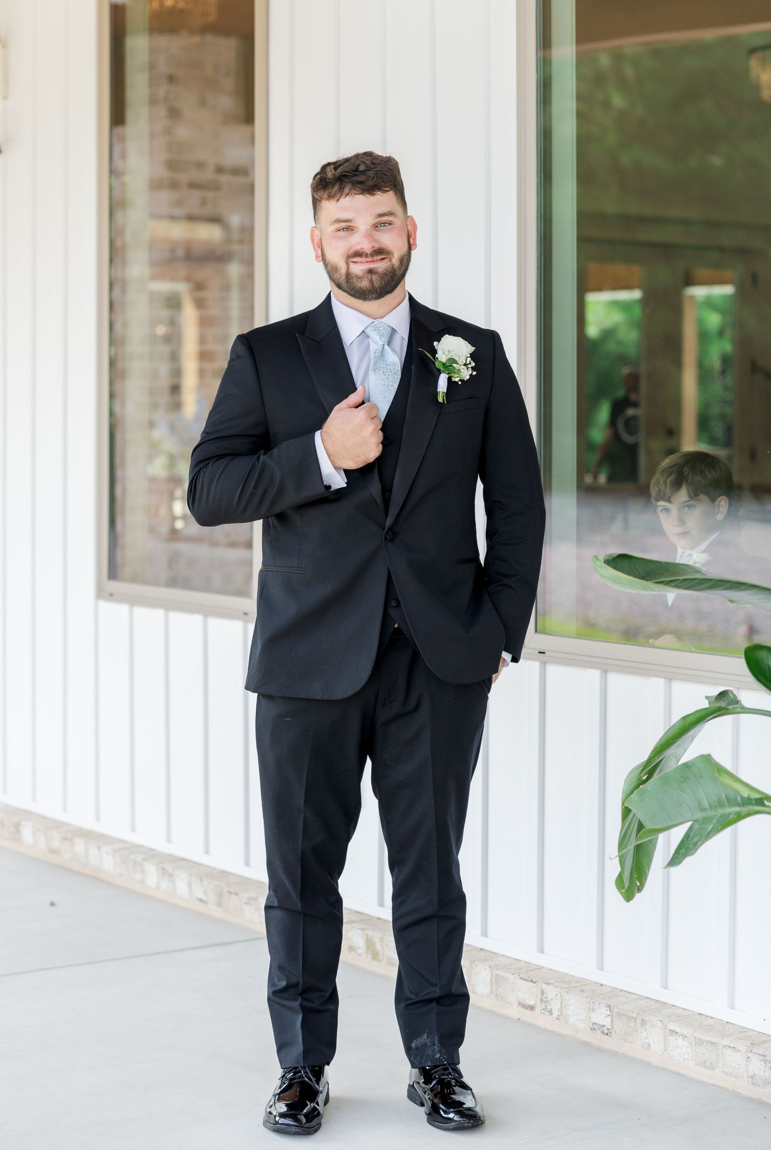 Handsome groom in black tuxedo with white boutonniere and light blue tie posing outdoors at wedding venue.