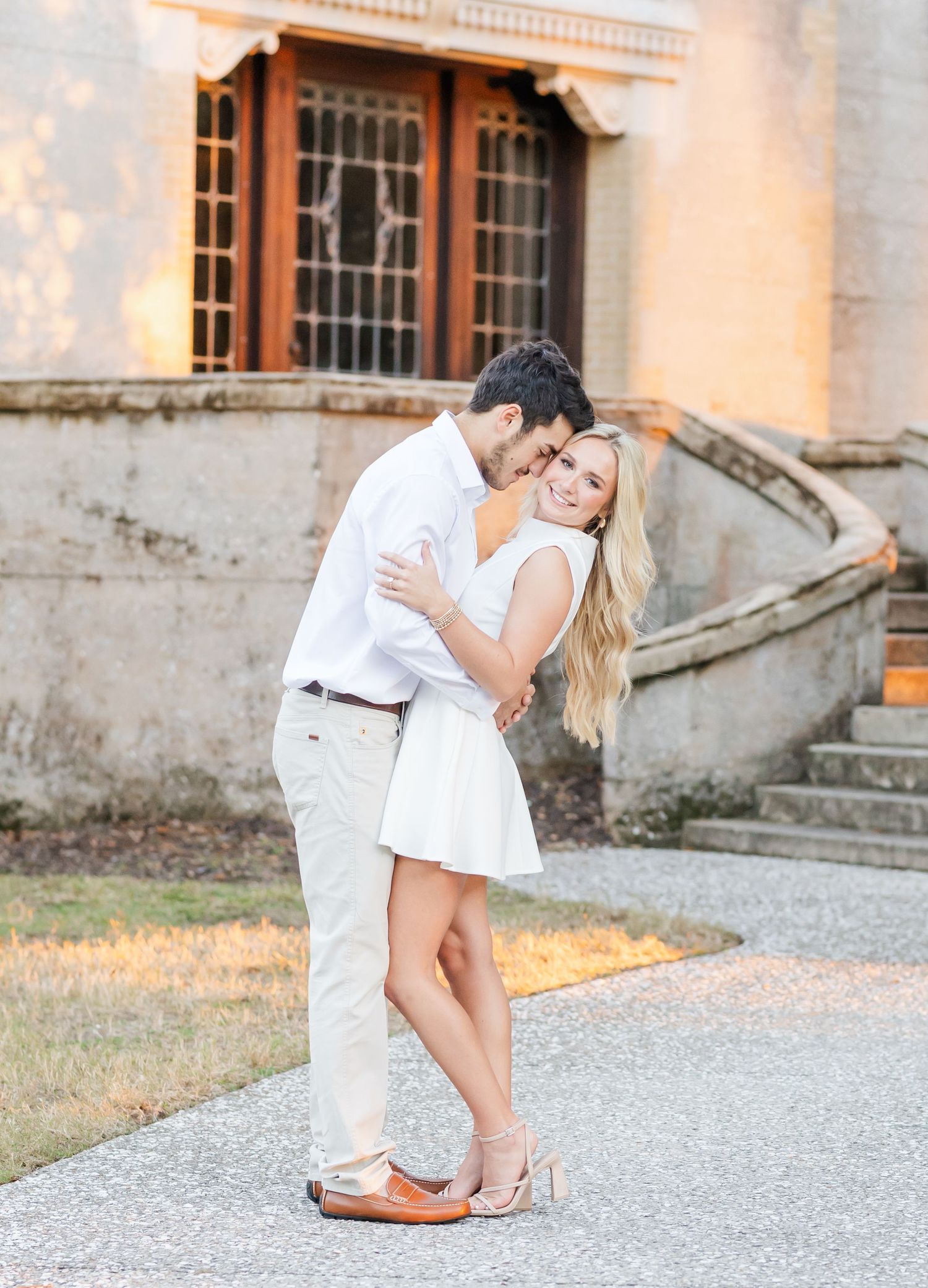 Romantic couple embracing on gravel path against historic stone building at golden hour.