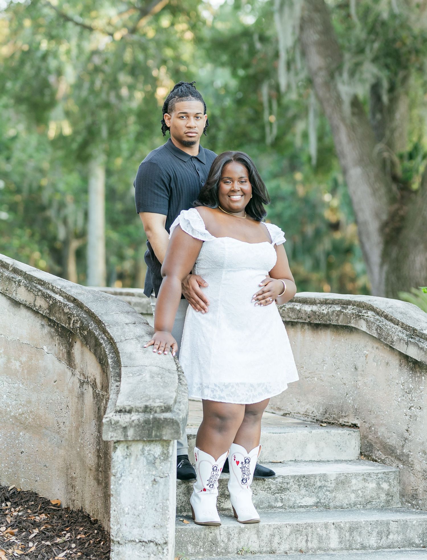 A couple in coordinating white and black outfits pose together on stone steps in a garden setting.