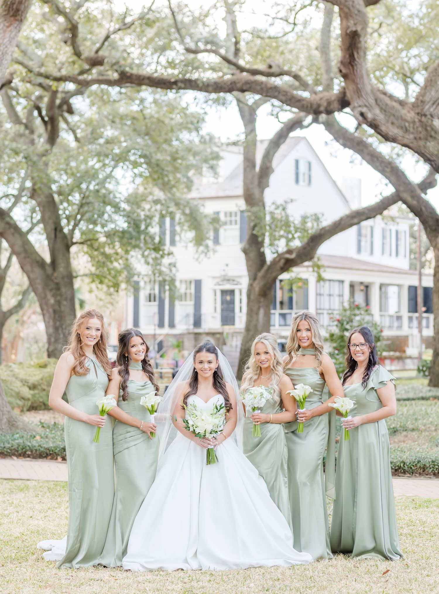 Wedding party in sage green bridesmaid dresses and white bridal gown posing under oak trees.