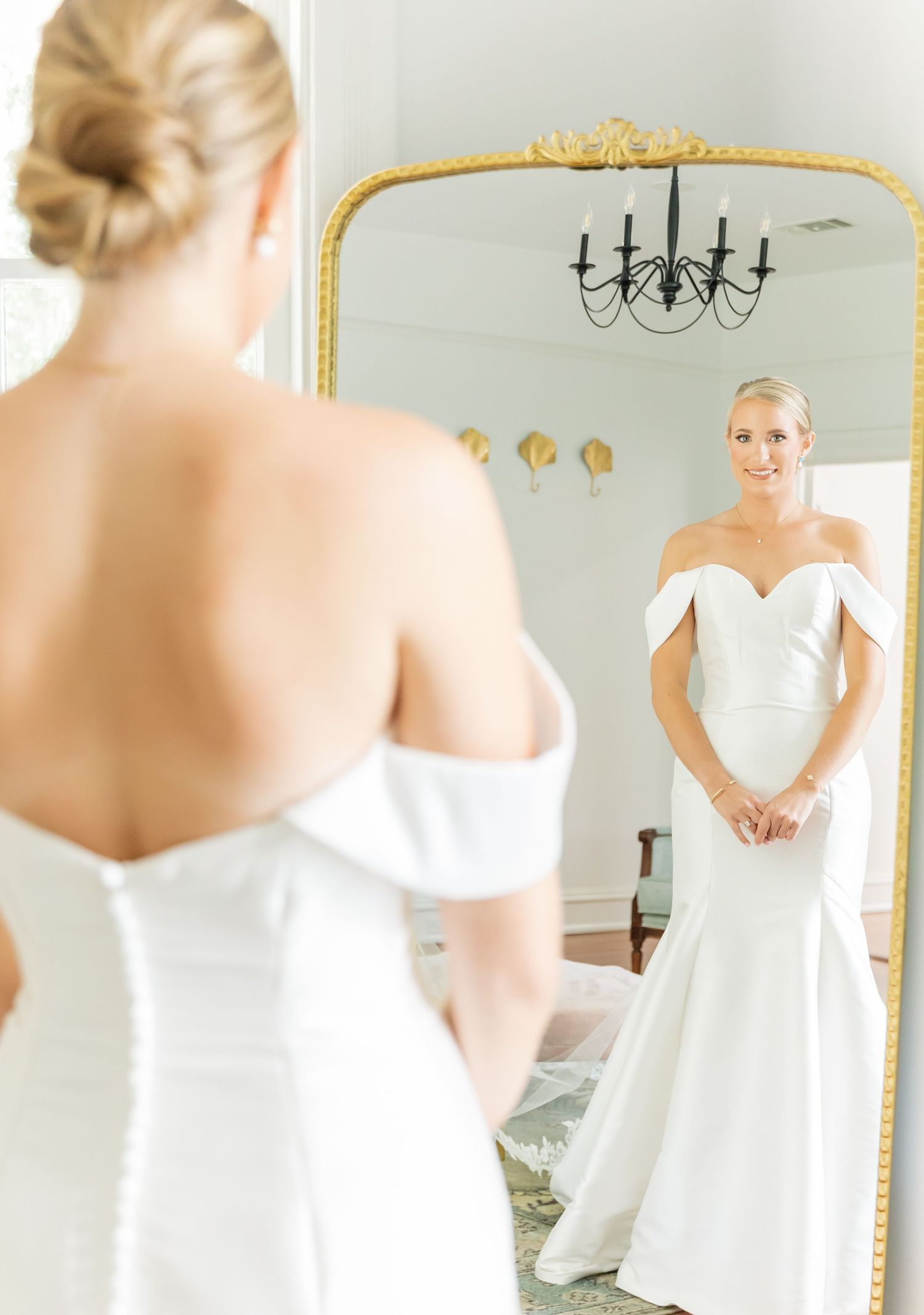 Elegant off-shoulder white wedding dress reflected in an ornate gold mirror with chandelier.