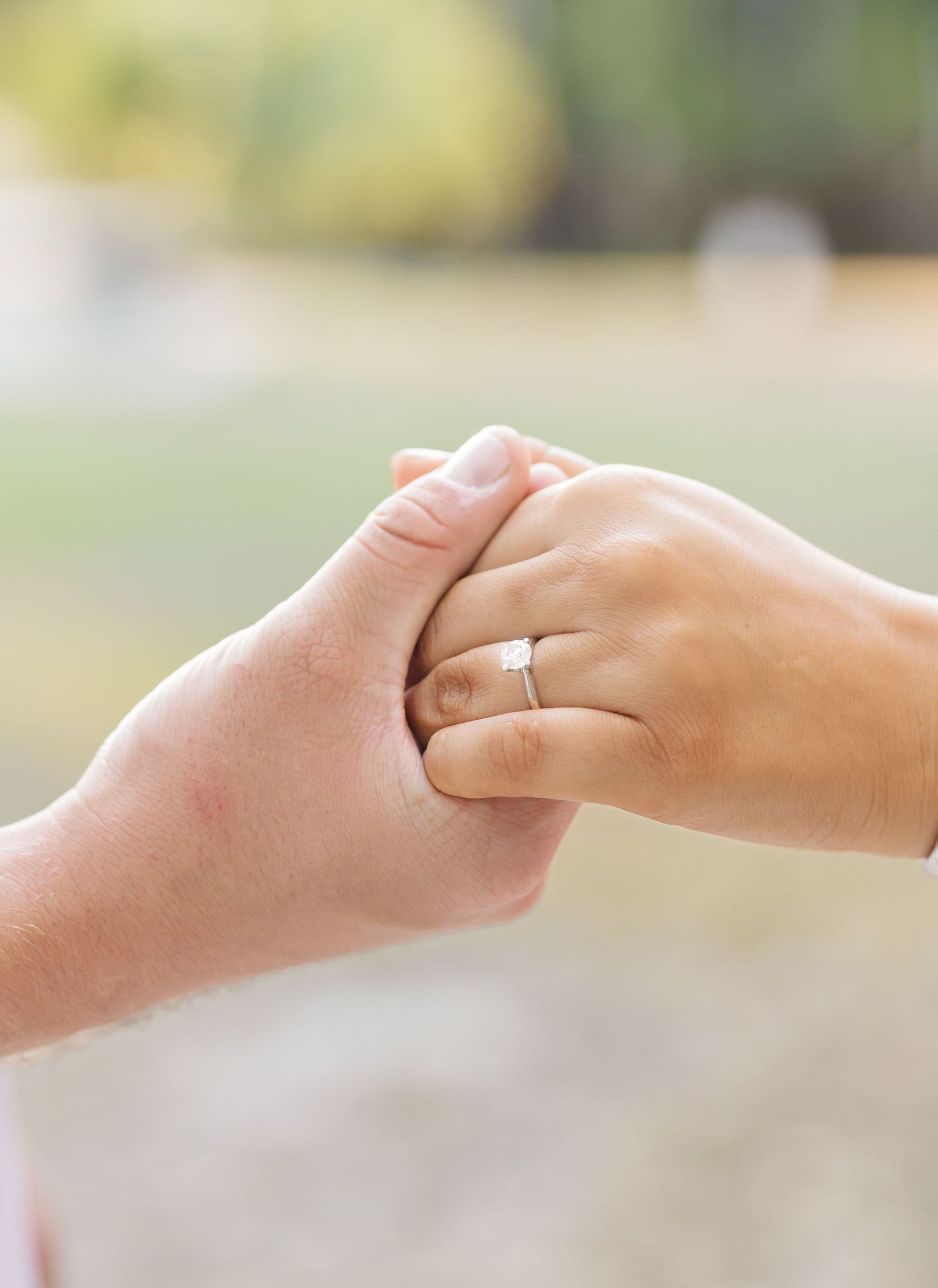 Two hands gently clasped together, with one wearing an engagement ring, in a romantic outdoor setting.