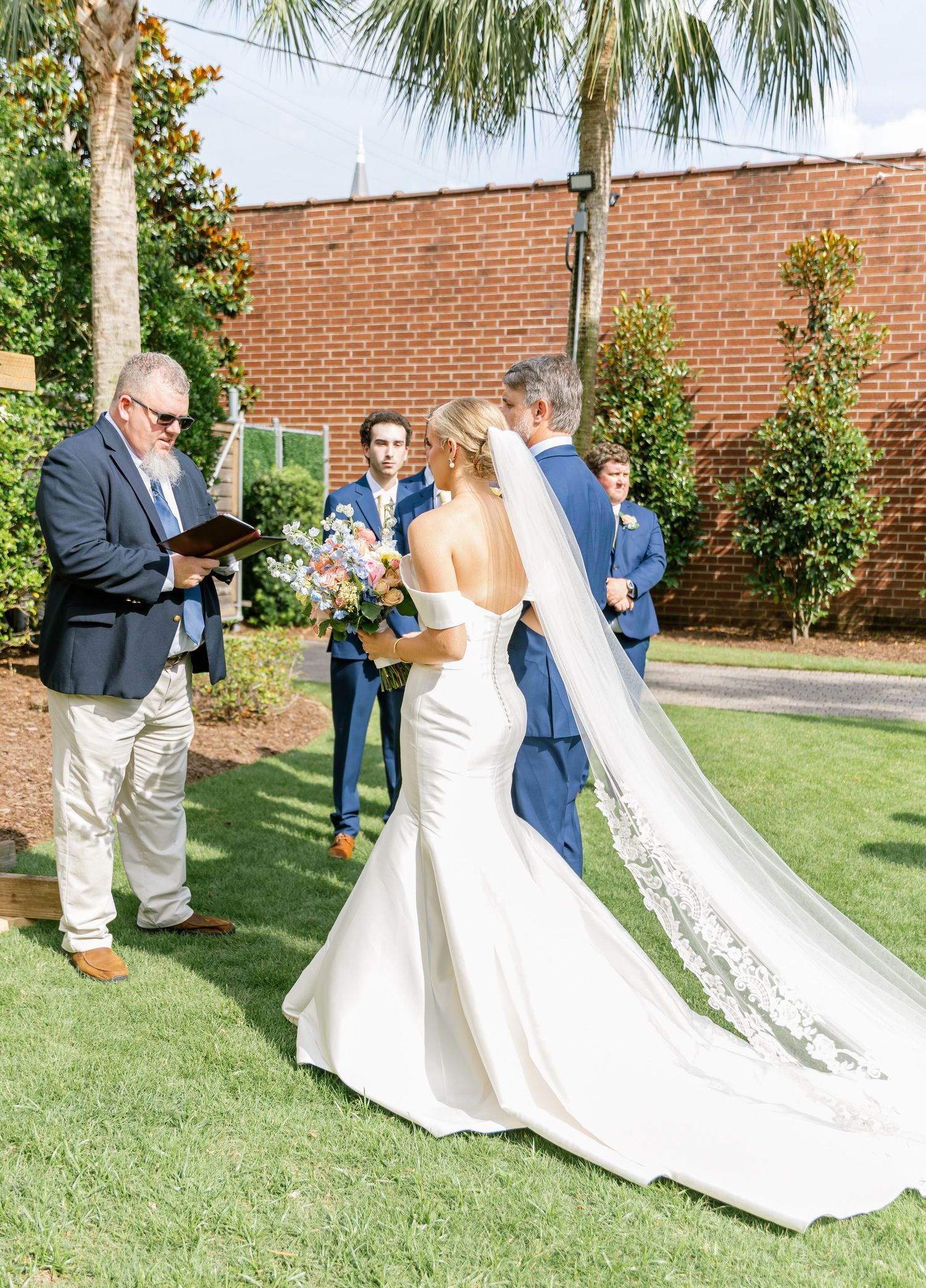 A wedding ceremony takes place outdoors with the couple and officiant standing on green grass near a brick wall.