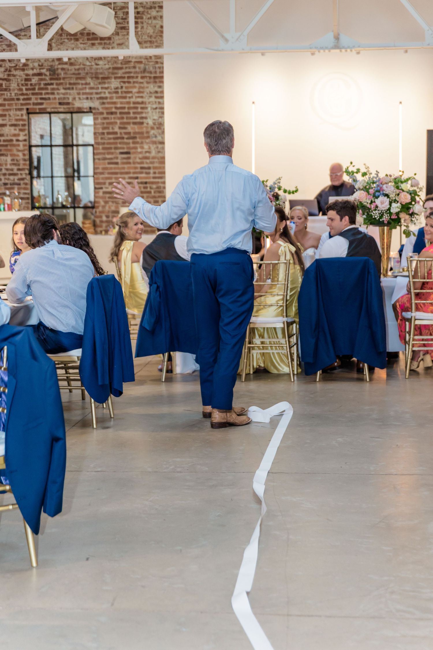 Wedding guests in navy blue attire dance in sequence across a spacious brick-walled reception venue.