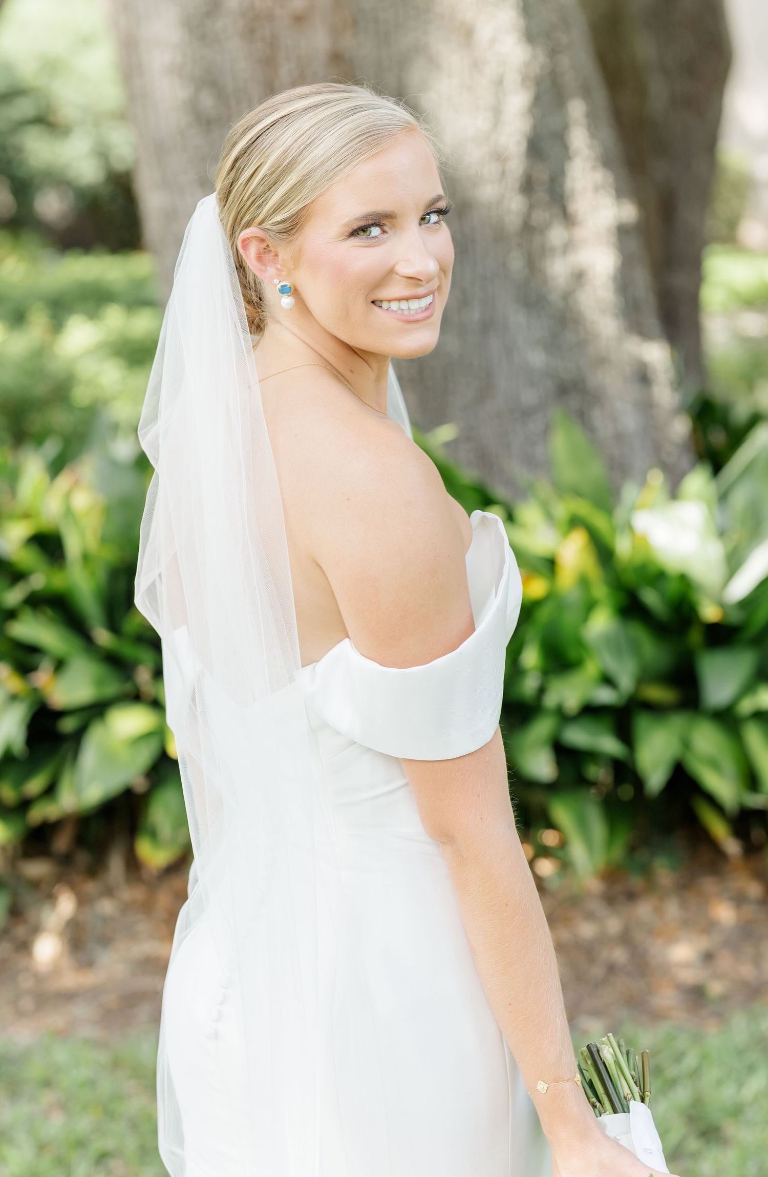 Elegant bride in white wedding dress and veil poses outdoors in a garden setting with natural greenery backdrop.