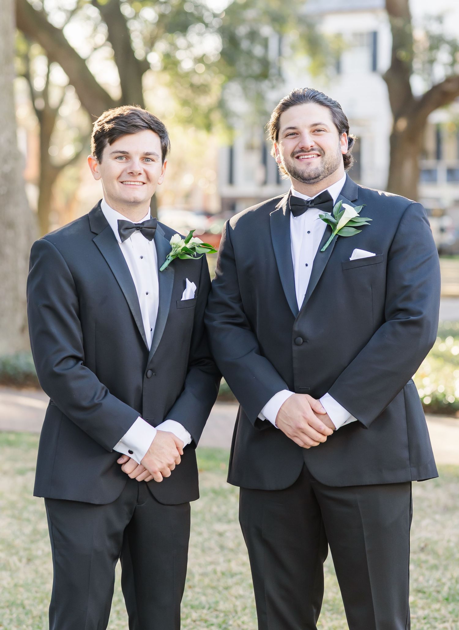 Wedding party groomsmen in formal black tuxedos pose casually outdoors.